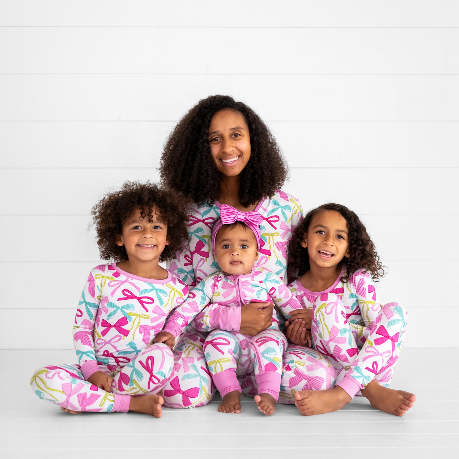 mom and her three children sitting together wearing matching Ribbons and Bows pj sets in women's and children's two piece and zippy styles. Her youngest is wearing a matching Pink Tonal Stripes luxe bow headband