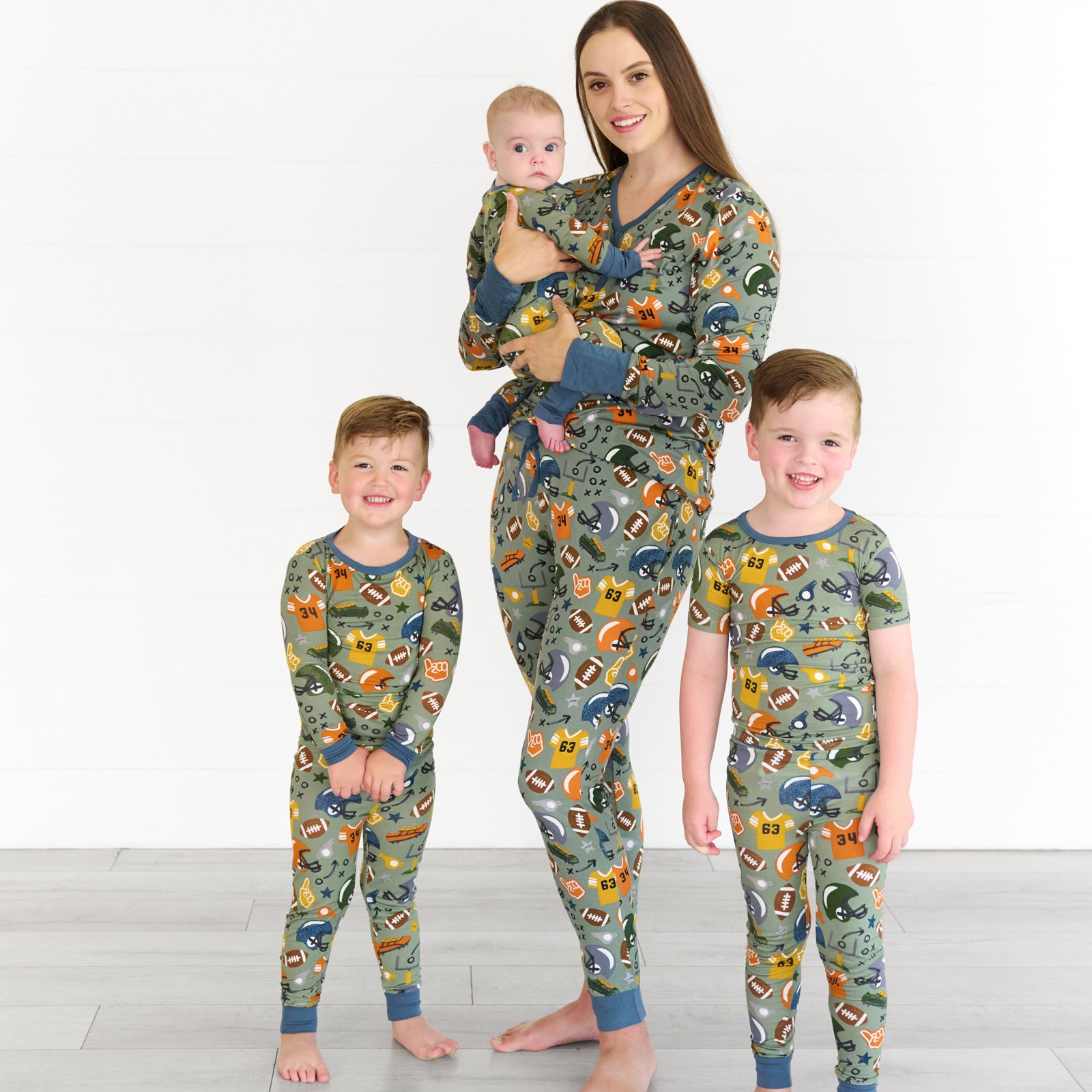 Boy on the left is wearing the touchdown-time-two-piece-pajama-set, female model in the middle is wearing the touchdown-time-womens-pajama-pants top, while holding baby in the touchdown-time-zippy. Boy on the right is the touchdown-time-two-piece-short-sleeve-pajama-set