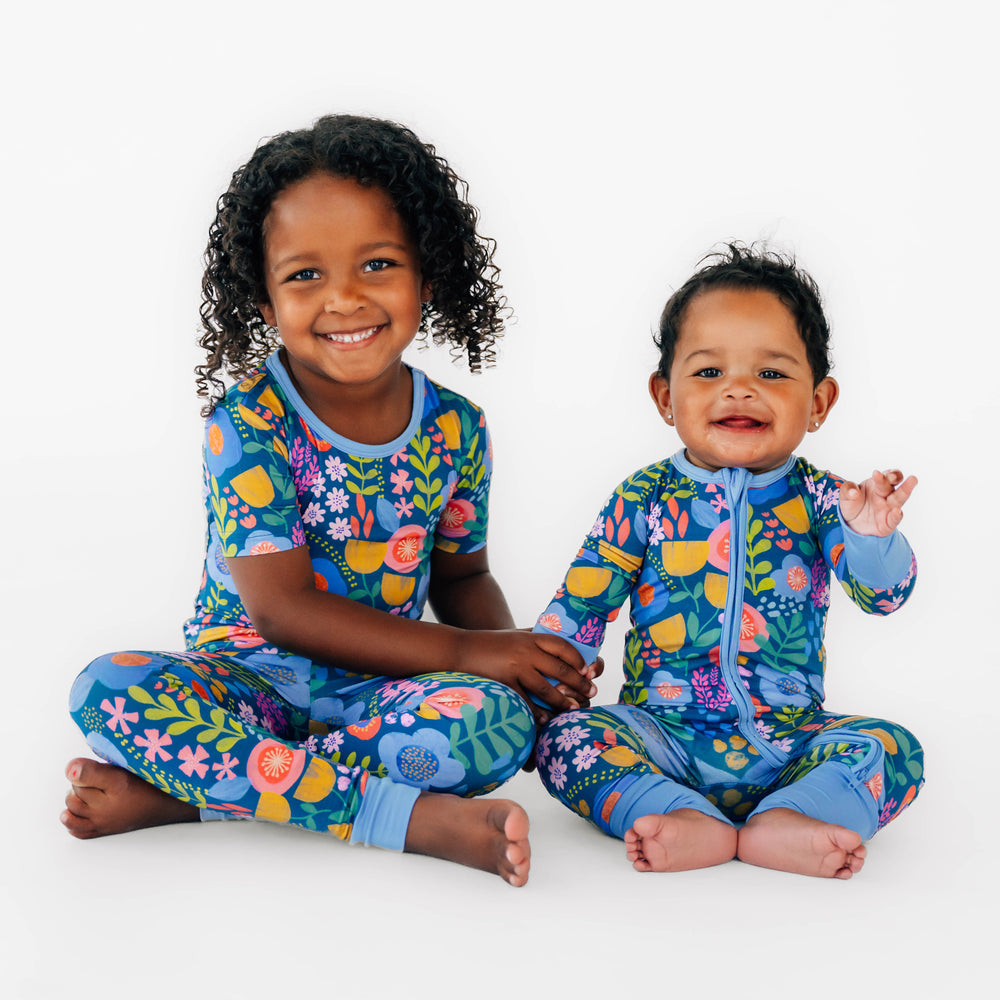 Two children sitting while wearing the Folk Floral. Girl on the left is wearing the Folk Floral Short Sleeve Pajama Set and baby on the left is wearing the Folk Floral Zippy