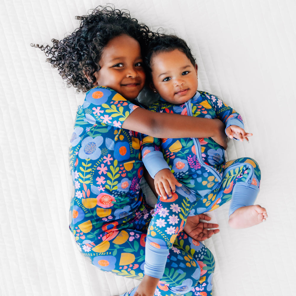 Two girls wearing the Folk Floral Print. Girl on left is wearing the Folk Floral Two-Piece Short Sleeve Pajama Set and baby on the right is wearing the Folk Floral Zippy