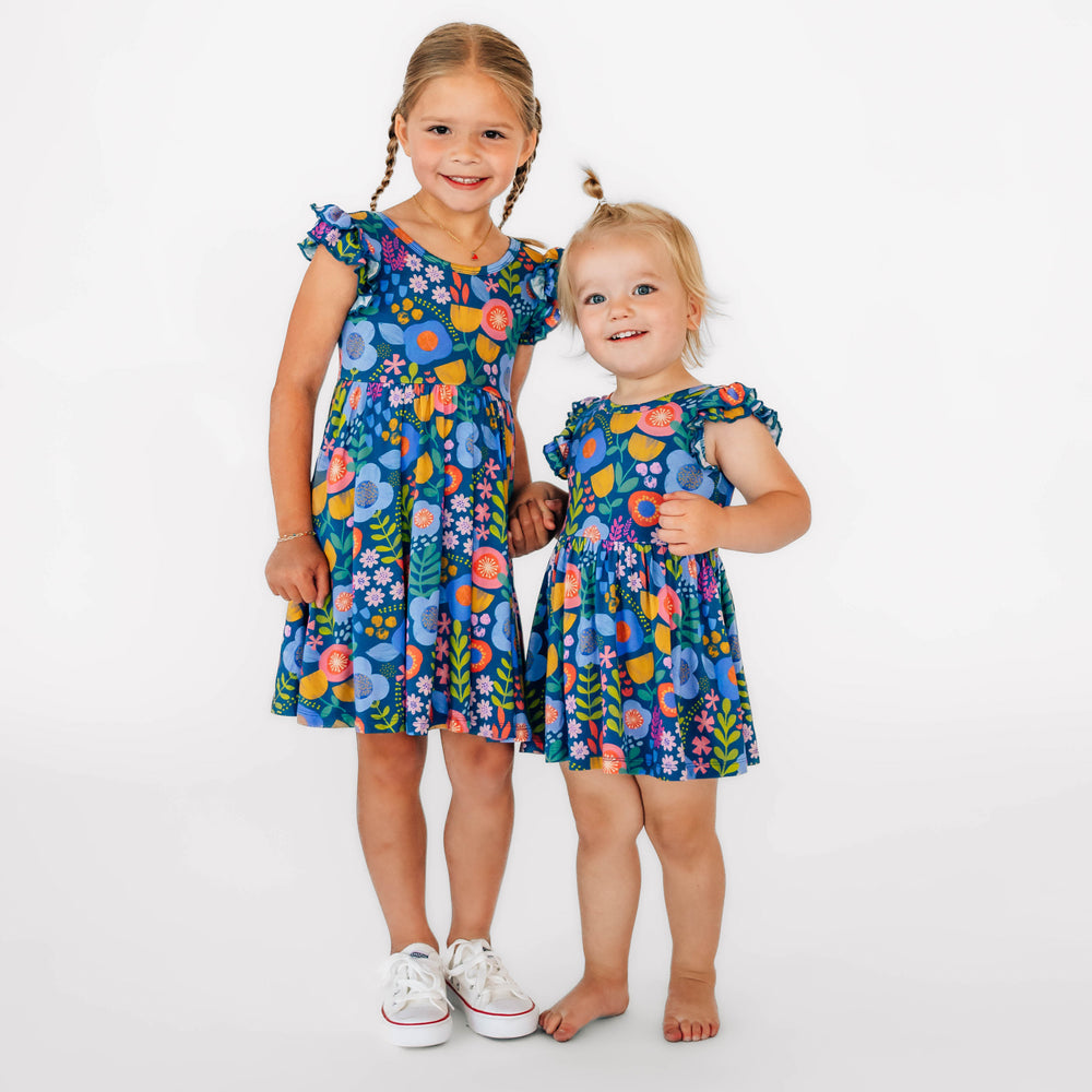 Two girls wearing the Folk Floral print. Girl on the right is wearing the Folk Floral Flutter Twirl Dress, and girl on the left is wearing the Folk Floral Flutter Twirl Dress with Bodysuit