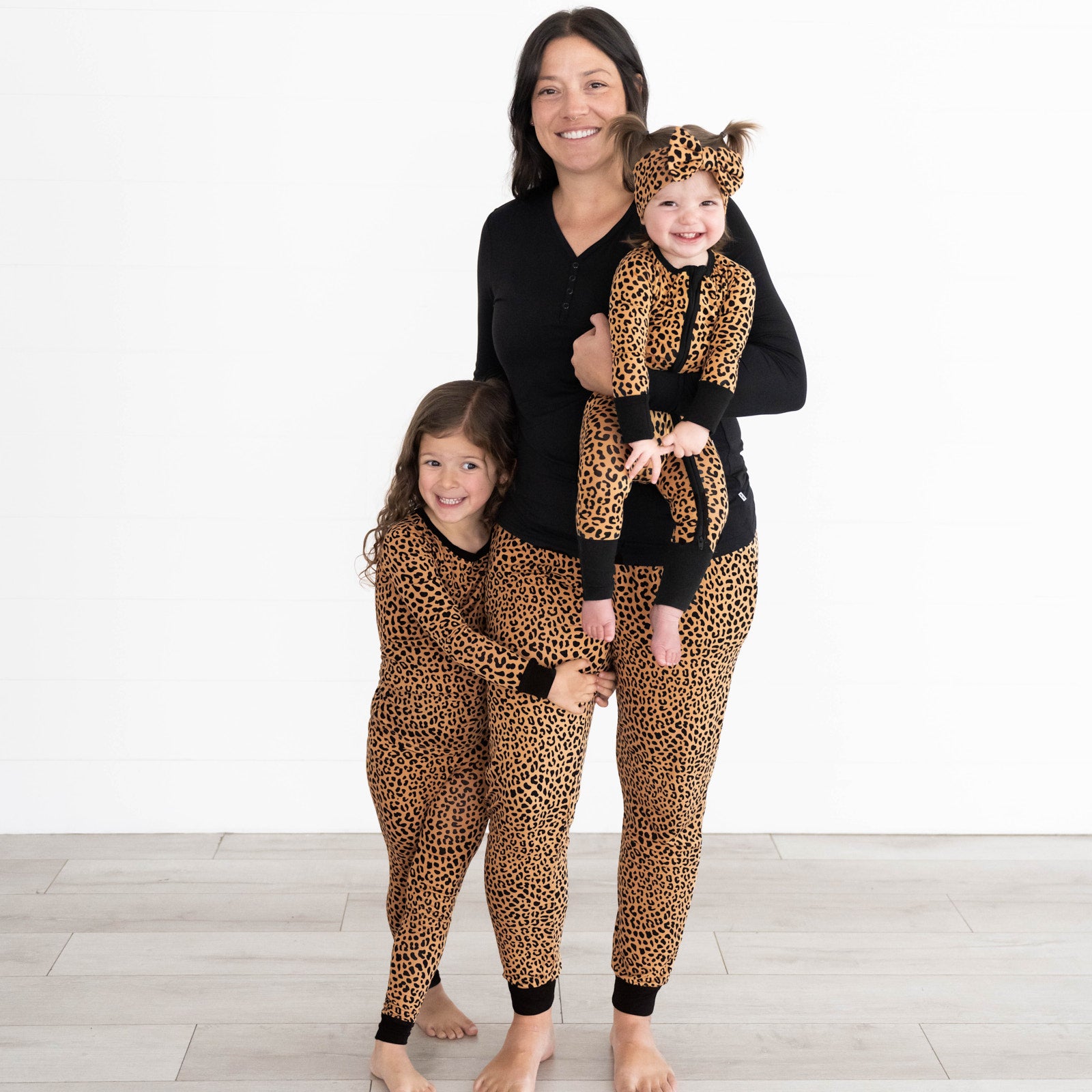 Girl on left is wearing the Classic Leopard Two-piece Pajama Set, mother in the middle is in the Classic Leopard Women's Pajama Pants & Black Women's Pajama Set. She is holding a baby wearing the the Classic Leopard Zippy and Luxe Bow