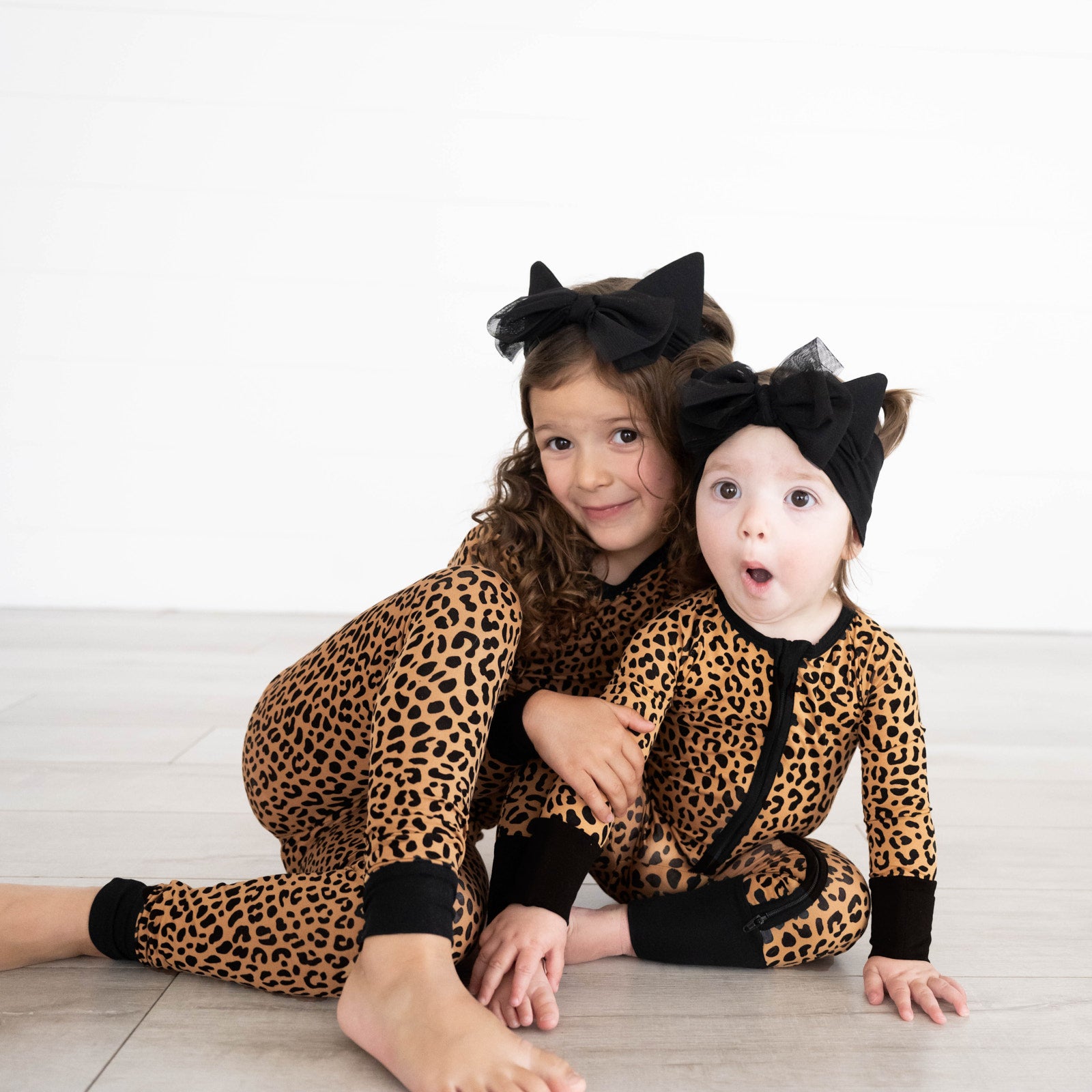 Girl on the left is wearing wearing the Classic Leopard Two-piece Pajama Set and girl on the right is wearing the Classic Leopard Zippy. Both are wearing the Car Ear Luxe Bow Headband