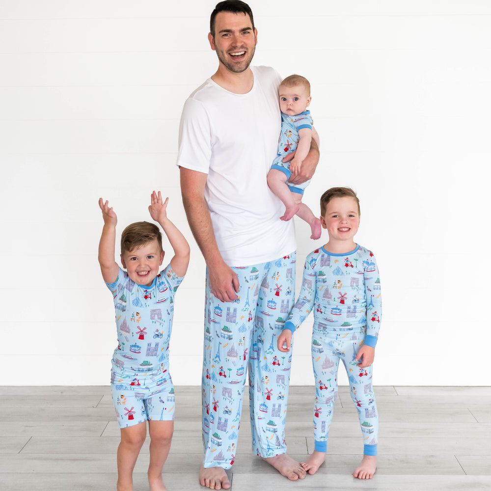Family of four wearing the Blue Weekend in Paris collection. Boy on the left is wearing the Blue Weekend in Paris Short Sleeve Pajama Set. Father in the middle is wearing the Blue Weekend in Paris Men's Pajama Pants while holding baby wearing the Blue Weekend in Paris Shorty Zippy. Boy on the right is wearing the Blue Weekend in Paris Pajama Set.