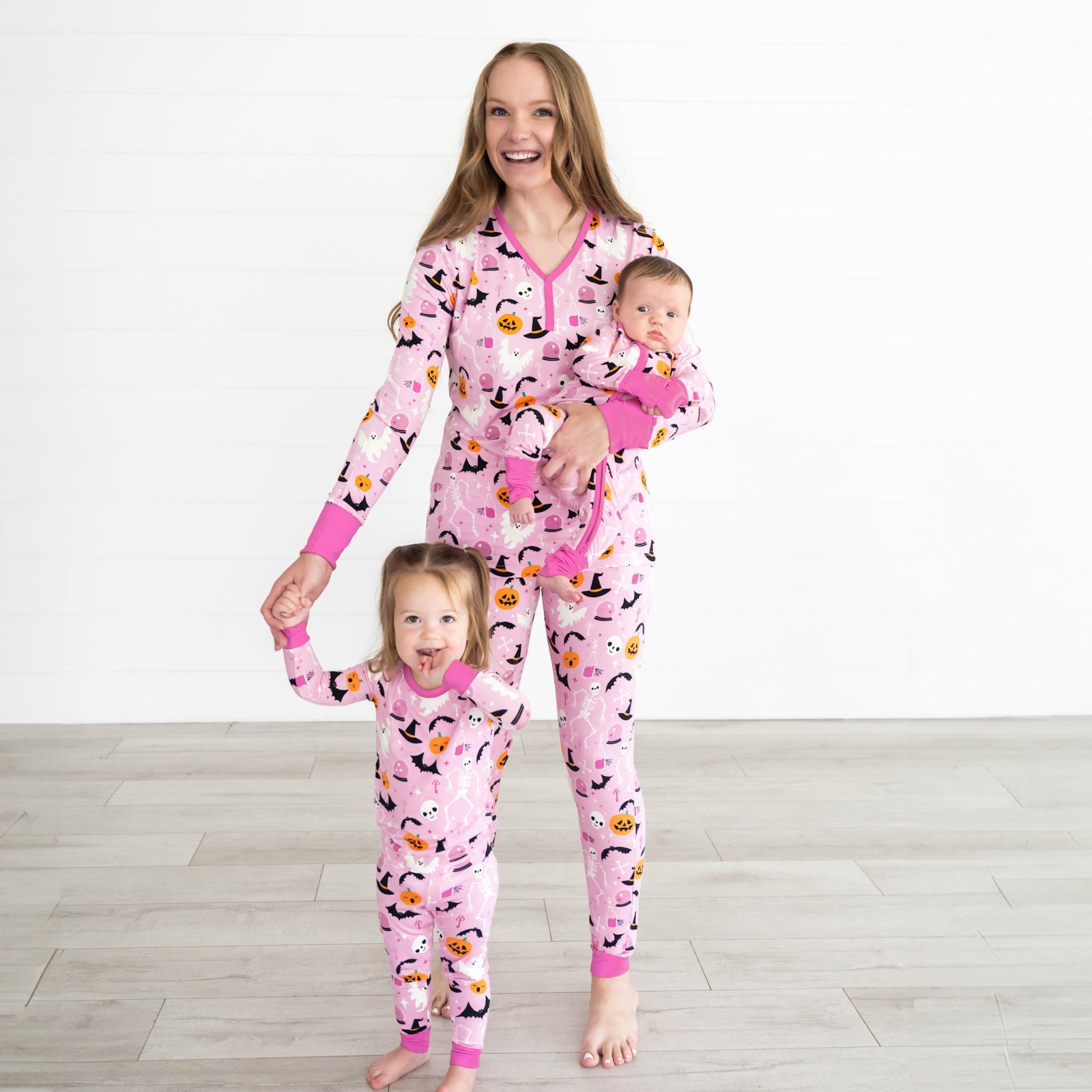 Girl on the left is wearing the Pink Glowing Ghouls Two-piece Pajama Set and mother is wearing the Pink Glowing Ghouls Women's Pajama Top & Pants. She is holding the baby, who is wearing the Pink Glowing Ghouls Zippy