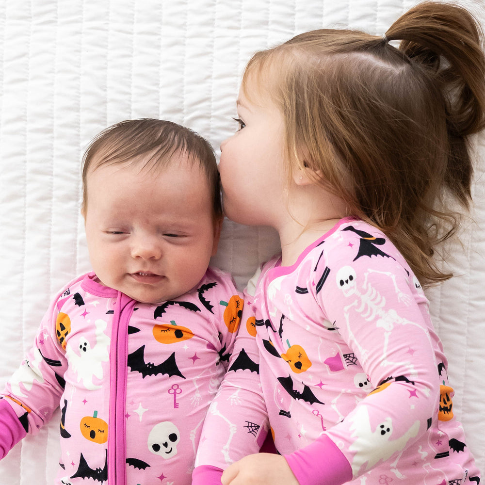 Baby on the left is wearing is laying down while wearing the Pink Glowing Ghouls Zippy and girl on the right is laying next to the baby while wearing the Pink Glowing Ghouls Two-piece Pajama Set