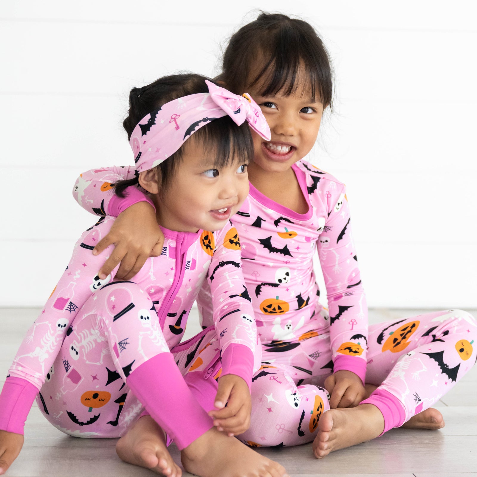 Two girls in the Pink Glowing Ghouls print. Girl on the left is in the Pink Glowing Ghouls Luxe Bow Headband and Pink Glowing Ghouls Zippy. Girl on the right is in the Pink Glowing Ghouls Two-piece Pajama Set