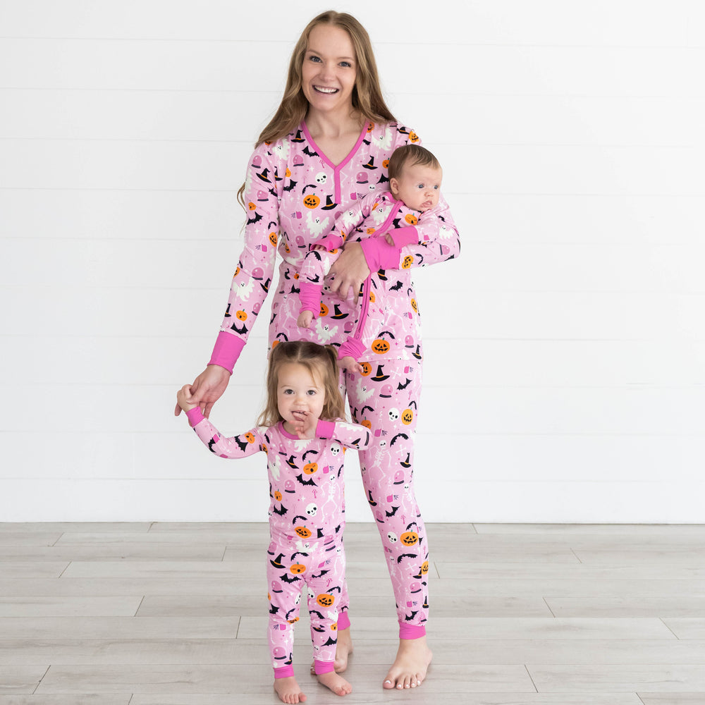 Girl in front is wearing the Pink Glowing Ghouls Two-Piece Pajama Set. Female model is wearing the Pink Glowing Ghouls Women's Top & Pants, while holding baby in the Pink Glowing Ghouls Zippy