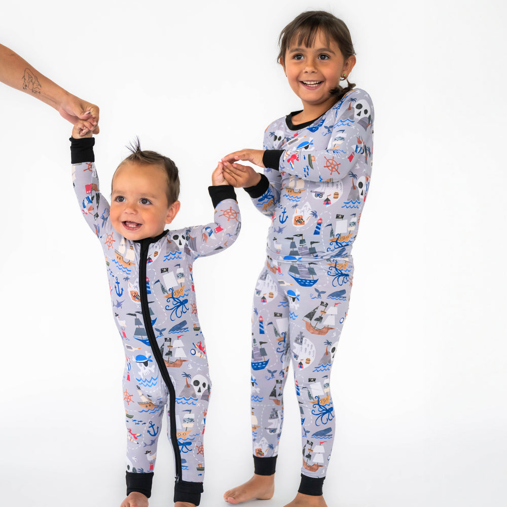 Girl and boy posing while wearing the Pirate's Map print. Boy on the left is wearing the Pirate's Map Zippy and girl is in the Pirate's Map Two-Piece Pajama Set
