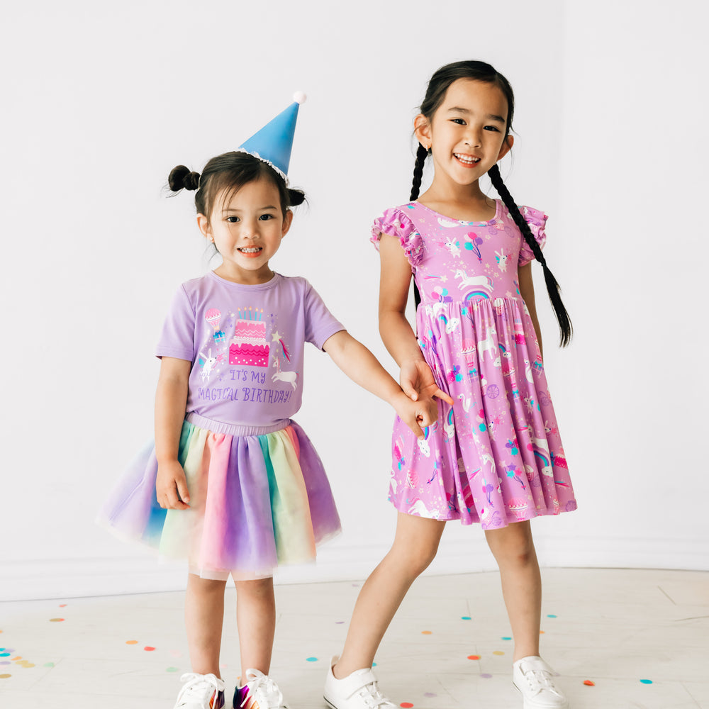 Child wearing a Magical Birthday flutter twirl dress holding hands with a child wearing a it's my birthday lavender tee paired with a rainbow tutu skirt