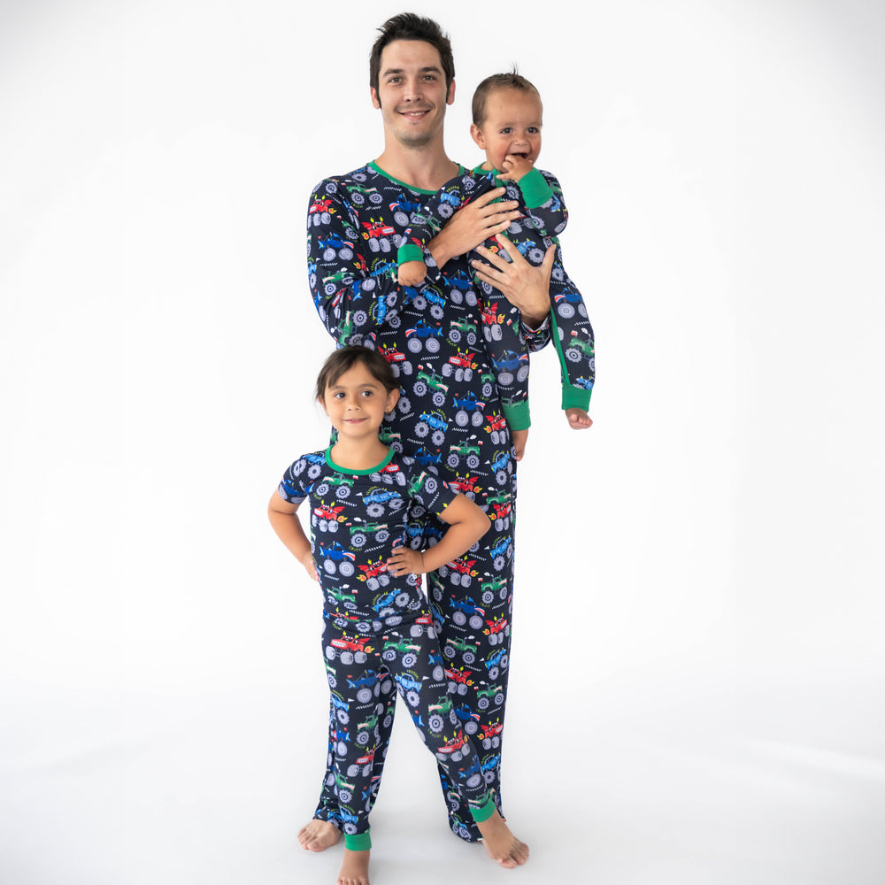 Girl on the left is posing in the Monster Truck Madness Two-Piece Short Sleeve Pajama Set. Father in the middle is wearing the Monster Truck Madness Men's Pajama Top & Pants, while holding the baby in a Monster Truck Madness Zippy