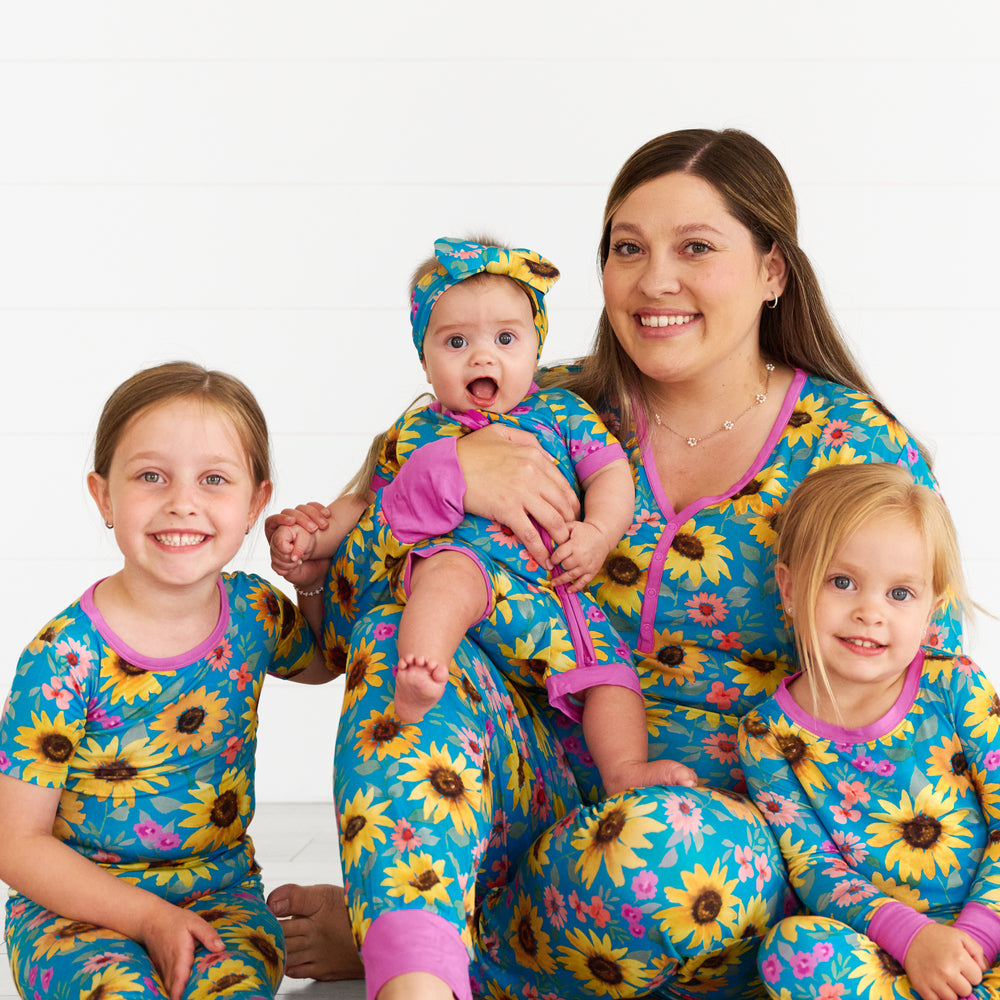 Girl on the left is wearing the Sunflower Fields Two-Piece Short Sleeve Pajama Set and holding baby's hand that is wearing the Sunflower Fields Shorty Zippy and Luxe Bow. Mother in the middle is in the Sunflower Fields Women's Pajama Pants and Top. Girl on the left is in the Sunflower Fields Two-Piece Pajama Set