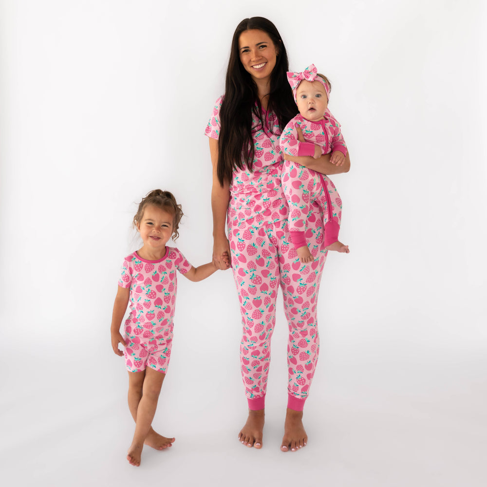 Mother and two daughters wearing the Sweet Strawberries print. Girl on the left is standing while wearing the Sweet Strawberries Two-Piece Short Sleeve & Shorts Pajama Set. The mother is wearing the Sweet Strawberries Women's Short Sleeve Pajama Top and Sweet Strawberries Women's Short Sleeve Pajama Pants, while holding the baby, who is wearing the Sweet Strawberries Zippy and Sweet Strawberries Luxe Bow Headband