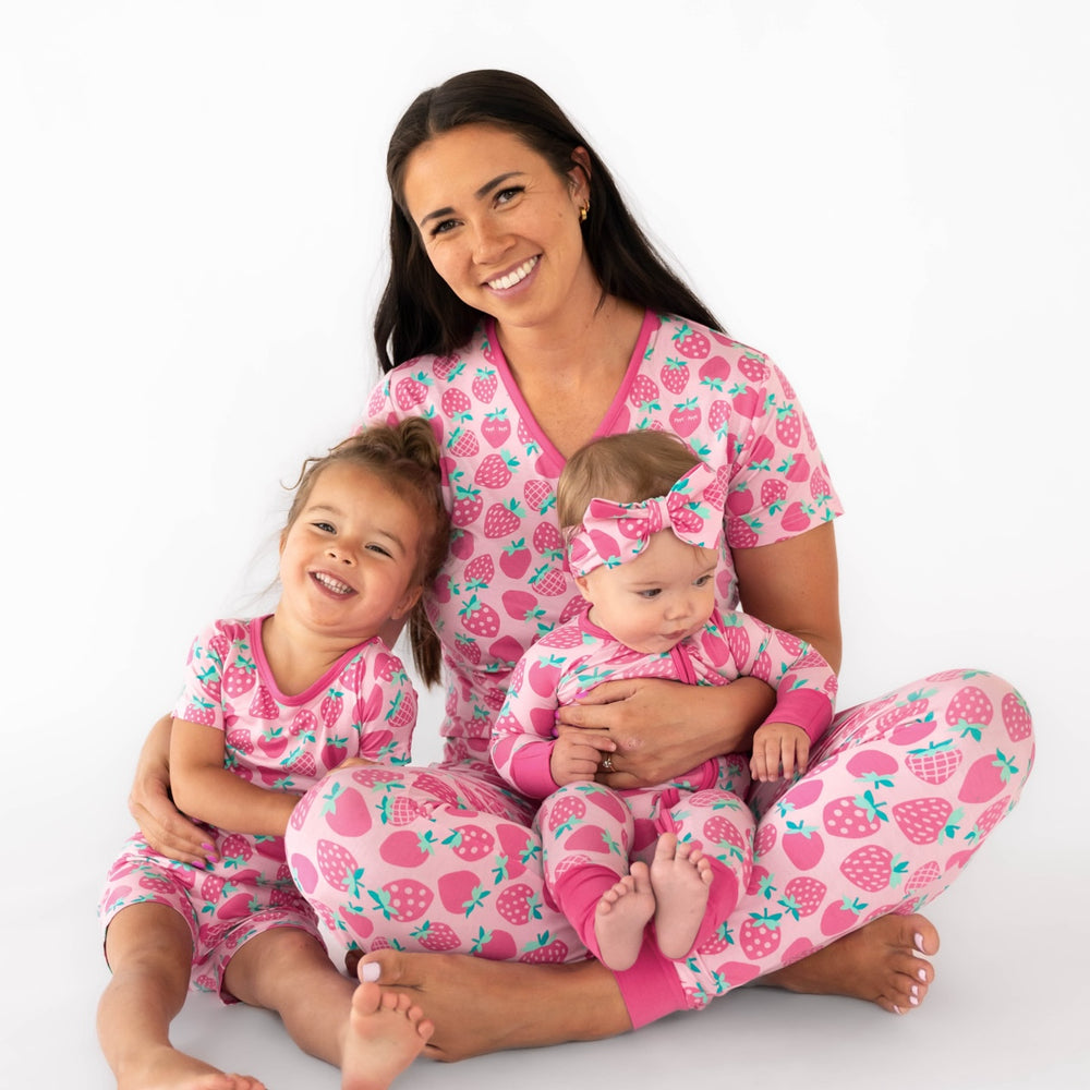 Mother and two daughters sitting while wearing the Sweet Strawberries print. Girl on the left is wearing the Sweet Strawberries Two-Piece Short Sleeve & Shorts Pajama Set. The mother is wearing the Sweet Strawberries Women's Short Sleeve Pajama Top and Pants, while holding the baby, who is wearing the Sweet Strawberries Zippy and Sweet Strawberries Luxe Bow Headband