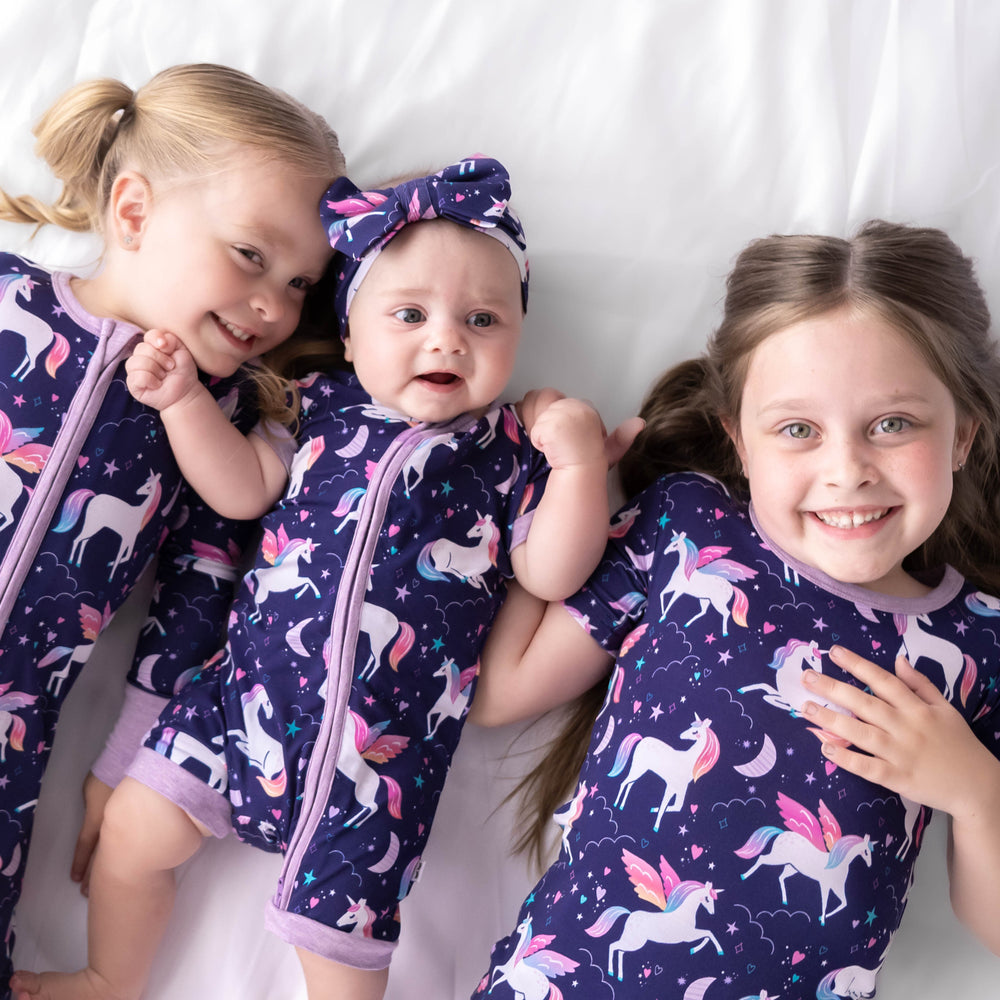 Three girl laying down while wearing the Magical Skies print. Girl on the left is wearing the Magical Skies Zippy and baby in the middle is wearing the Magical Skies Shorty Zippy. Girl on the right is wearing the Magical Skies Two-Piece Short Sleeve & Shorts Pajama Set