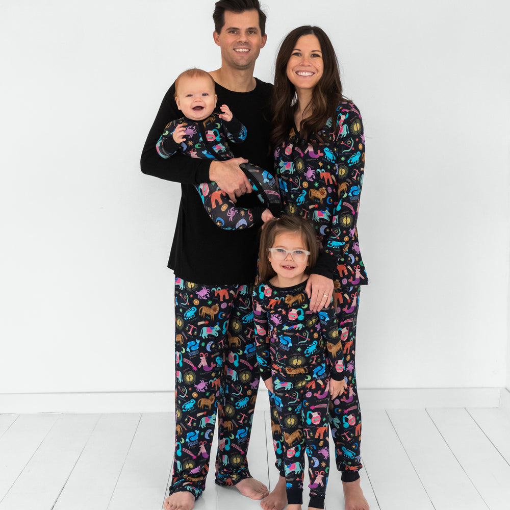 Family of four wearing matching Star Signs pjs. Mom is wearing women's Star Signs pj top and coordinating women's pj pants. Dad is wearing a men's black pj top and men's Star Signs pj pants. Their children are wearing a Star Signs zippy and matching two piece pj set.