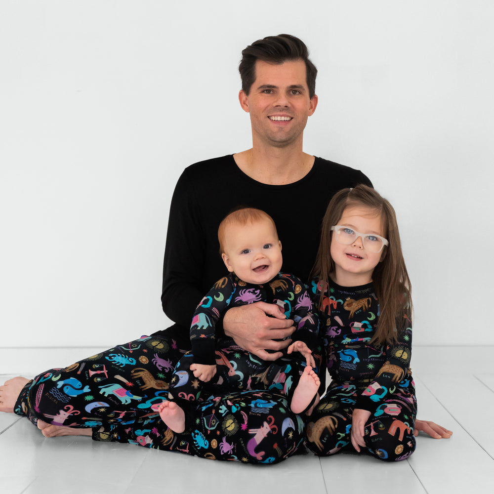 Dad and kids in coordinating in Star Signs printed pajama sets. Dad is wearing men's  Star Signs pajama pants and matching men's pajama top. Kids are wearing Star Signs two piece pajama set and a Star Signs zippy.