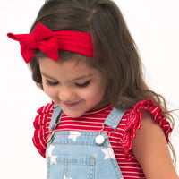 Close up image of a child wearing a Candy Red Stripes flutter tee and coordinating Play overalls and luxe bow headband