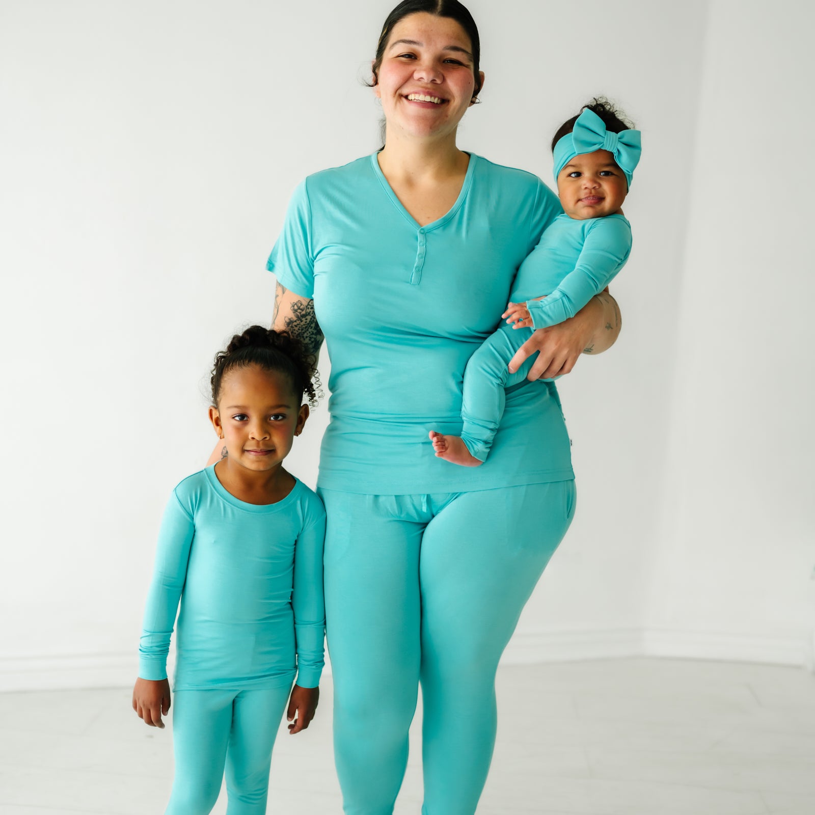 A mother and her two children wearing matching Glacier Turquoise pajamas. Mom is wearing women's Glacier Turquoise pajama top and matching women's pajama pants. Her children are wearing Glacier Turquoise pajamas in two piece and zippy styles