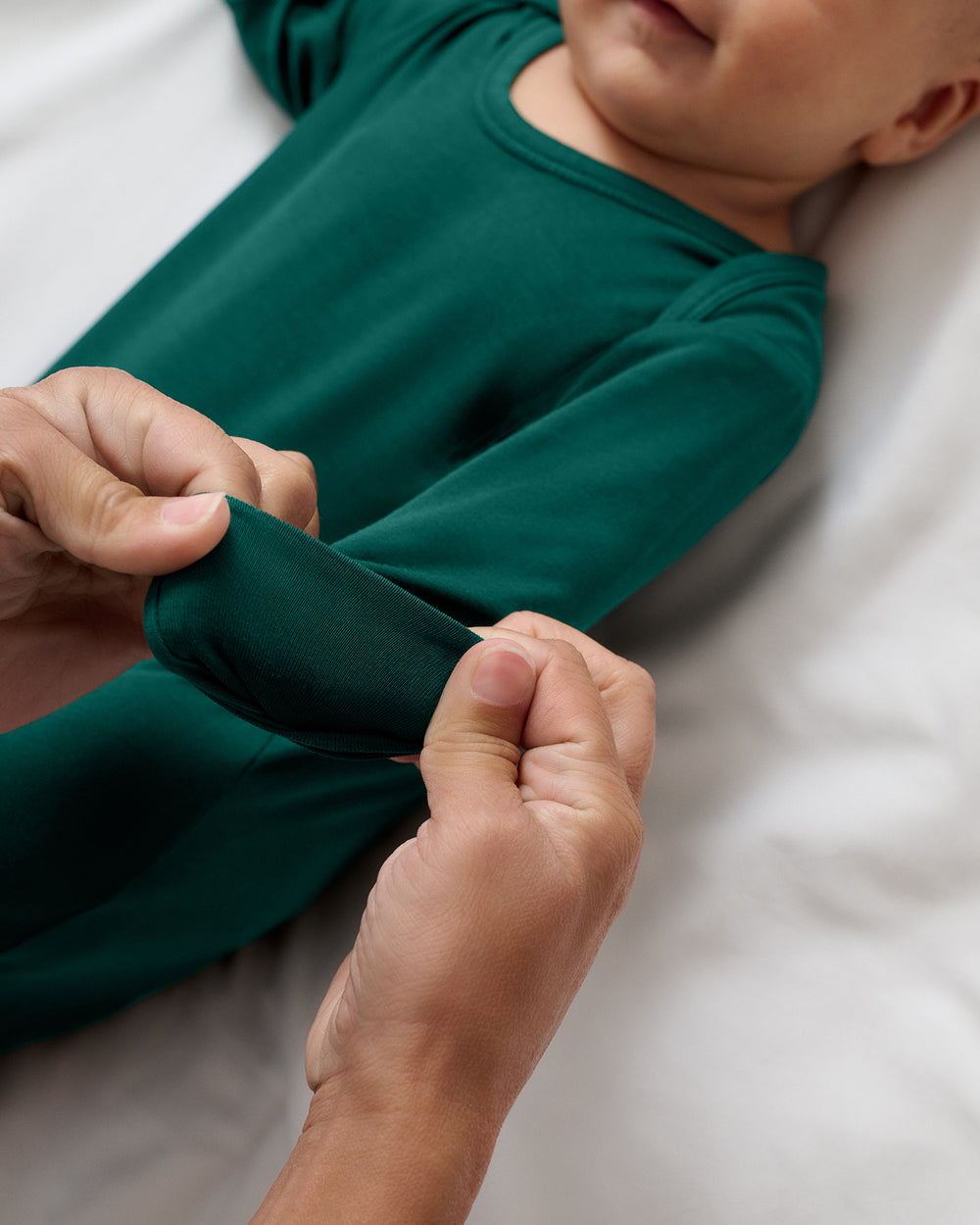 A close-up of a baby wearing a dark green long-sleeve one-piece gown, with an adult's hands stretching the fabric of the sleeve to show the mitten feature.