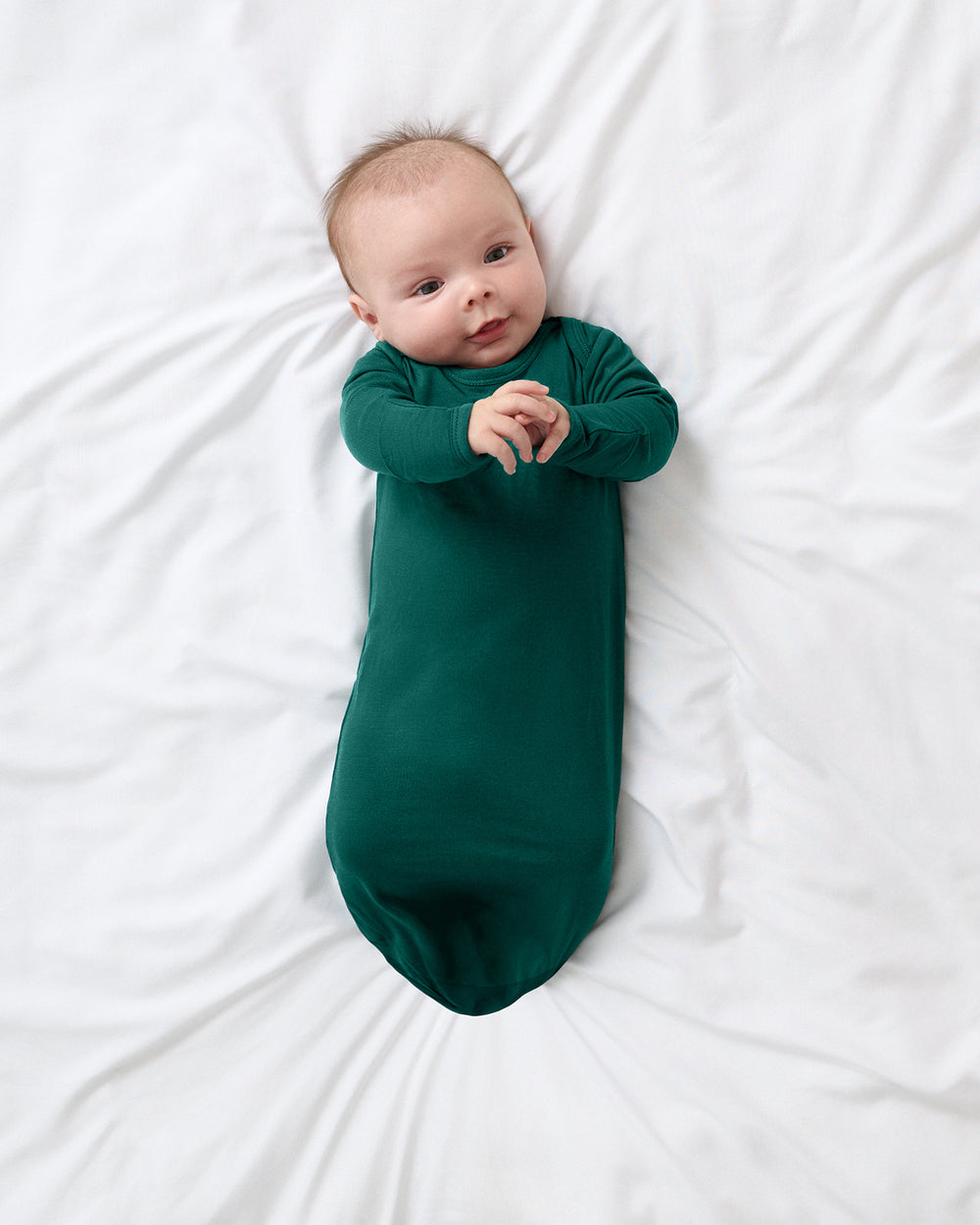 A baby lying on white bedding, dressed in a long-sleeve dark green sleep gown.