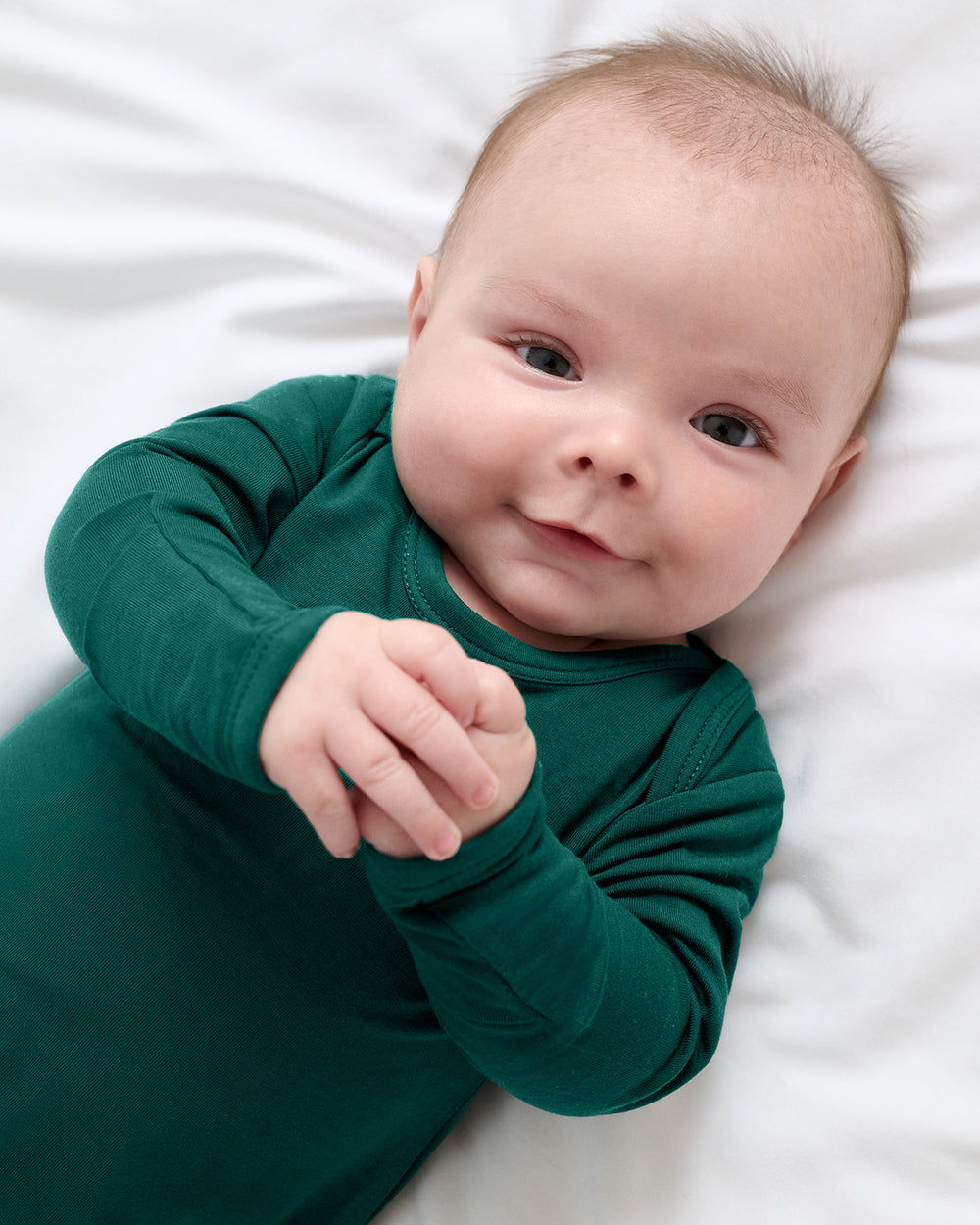A baby lying on white bedding, wearing a dark green long-sleeve sleep gown.