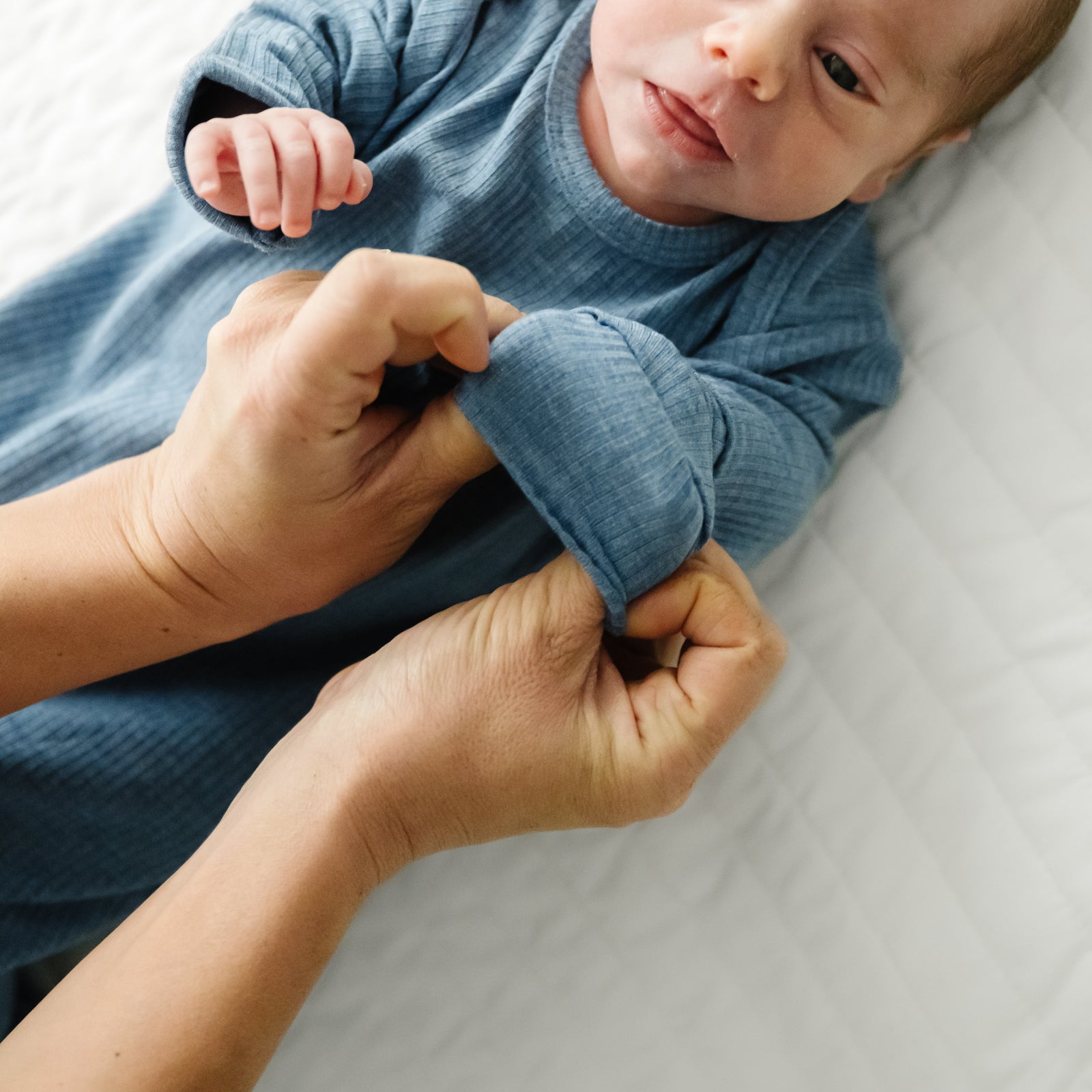 Child laying on a bed wearing a Heather Blue Ribbed Infant Gown demonstrating the fold over mittens