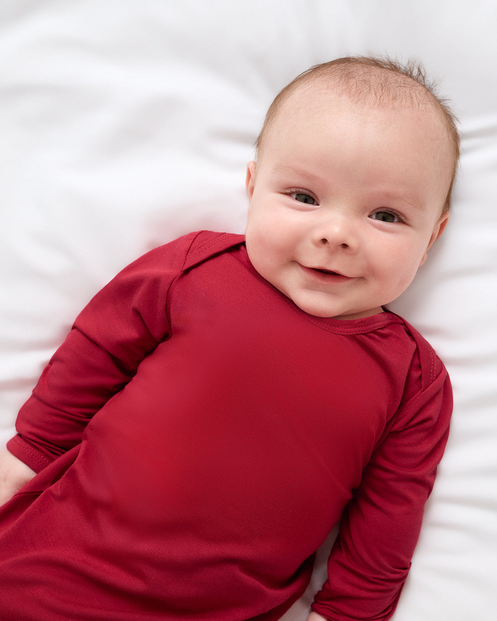 A smiling baby lying on a white blanket, wearing a solid deep red long-sleeve sleep gown.