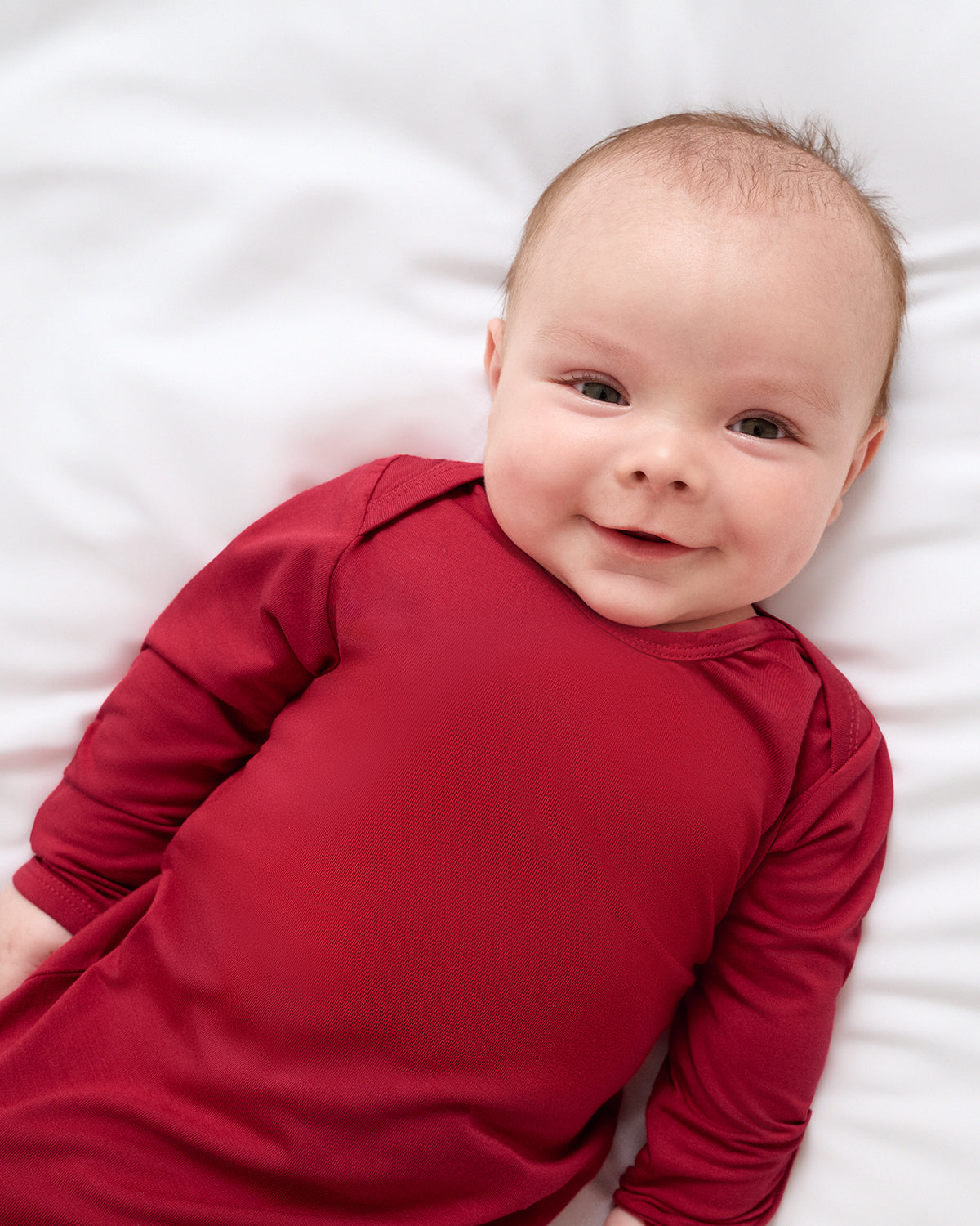 A smiling baby lying on a white blanket, wearing a solid deep red long-sleeve sleep gown.