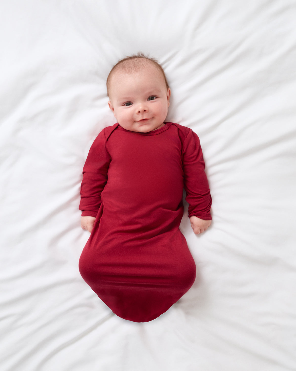 A baby lying on a white sheet, wearing a long-sleeve, footless red sleep sack.