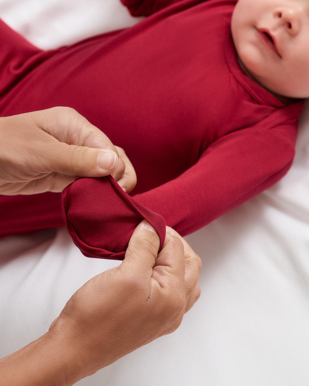 A close-up of a baby wearing a long-sleeve red gown, with an adult's hands adjusting the cuff of the sleeve on a white background.