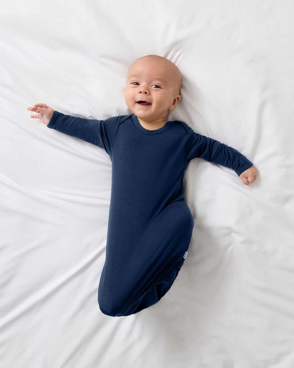 A smiling baby lying on a white bed, wearing a Classic Navy Infant Gown.