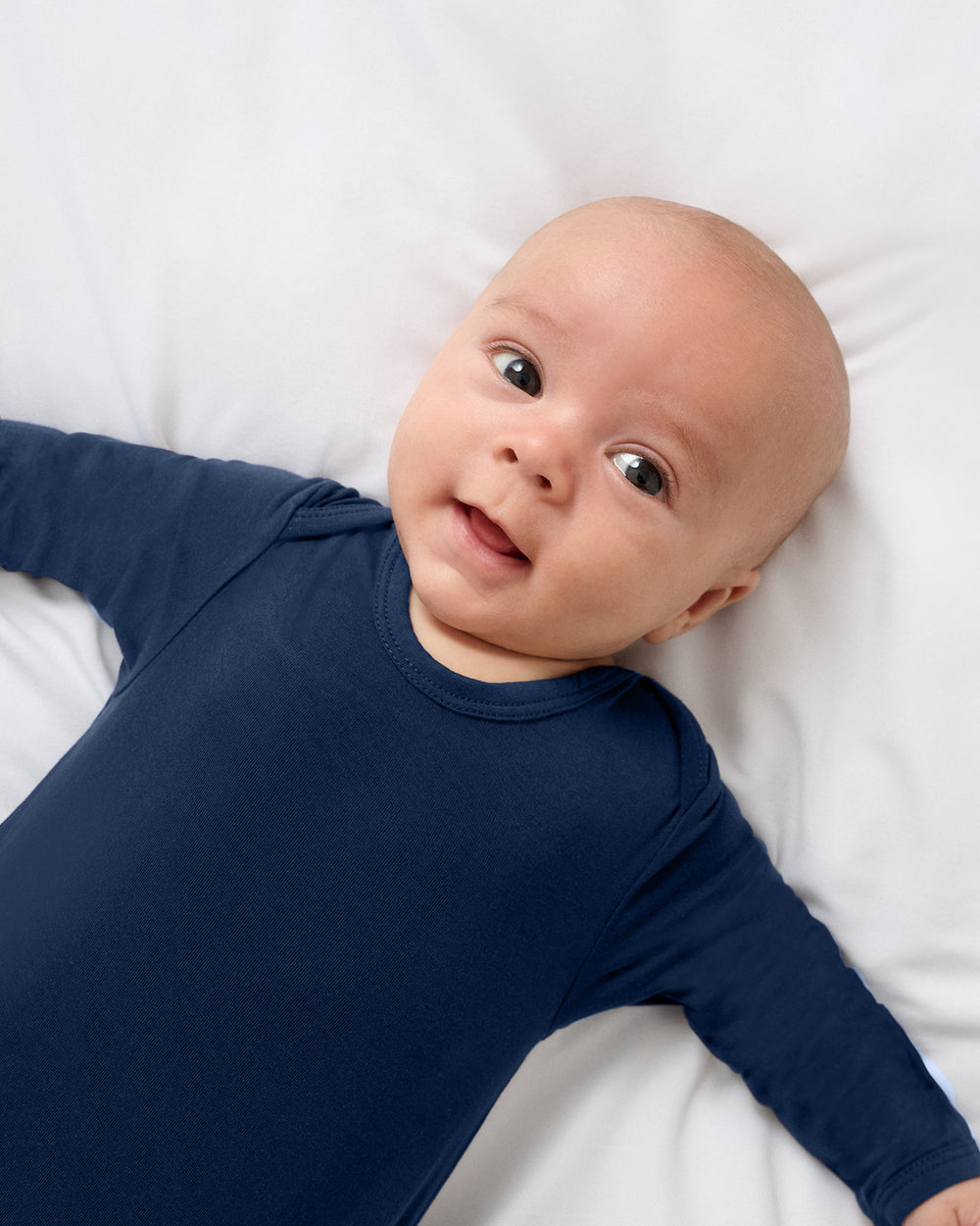 A baby lying on a white blanket, wearing a solid Classic Navy Infant Gown.