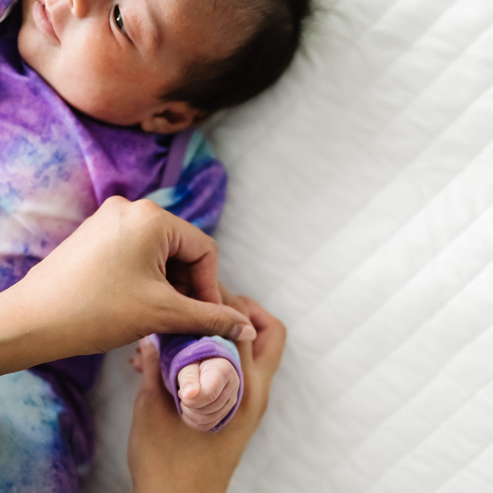 Child laying on a bed wearing a Purple Watercolor Infant Gown demonstrating the fold over mittens
