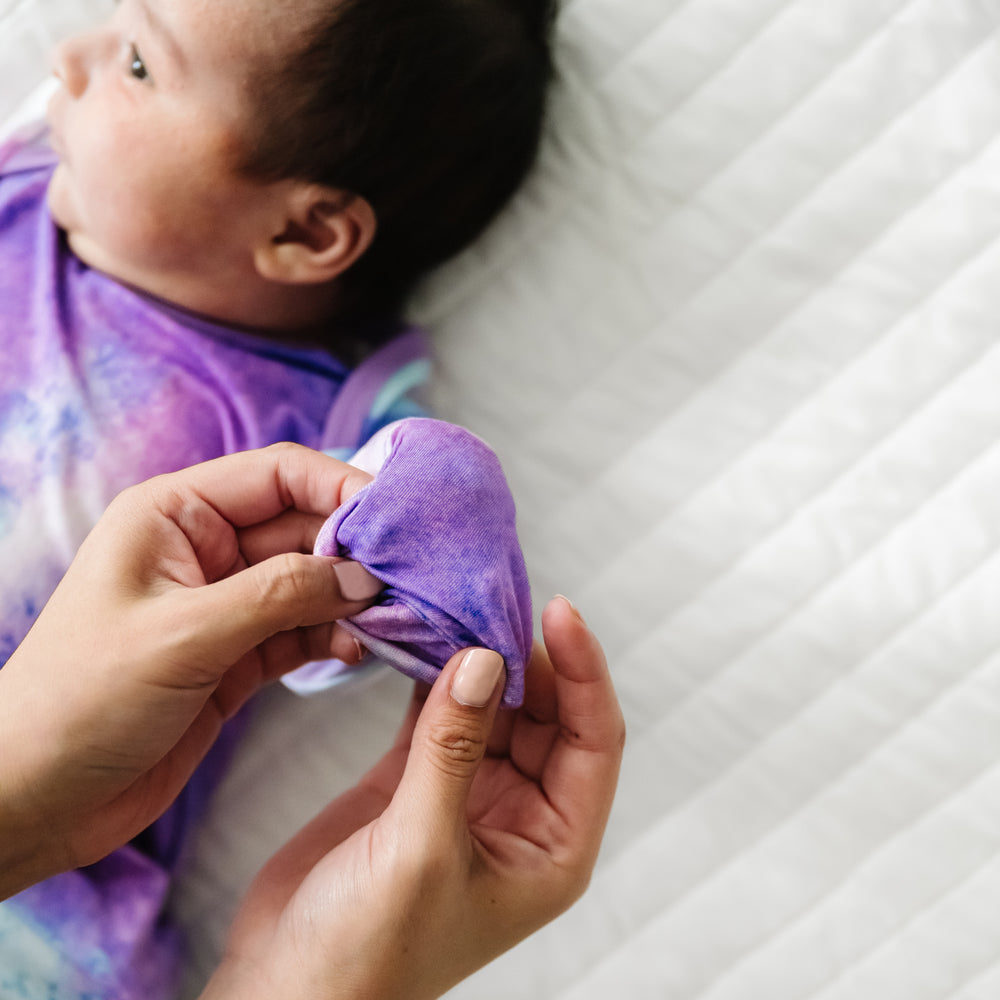 Alternate image of a child laying on a bed wearing a Purple Watercolor Infant Gown demonstrating the fold over mittens