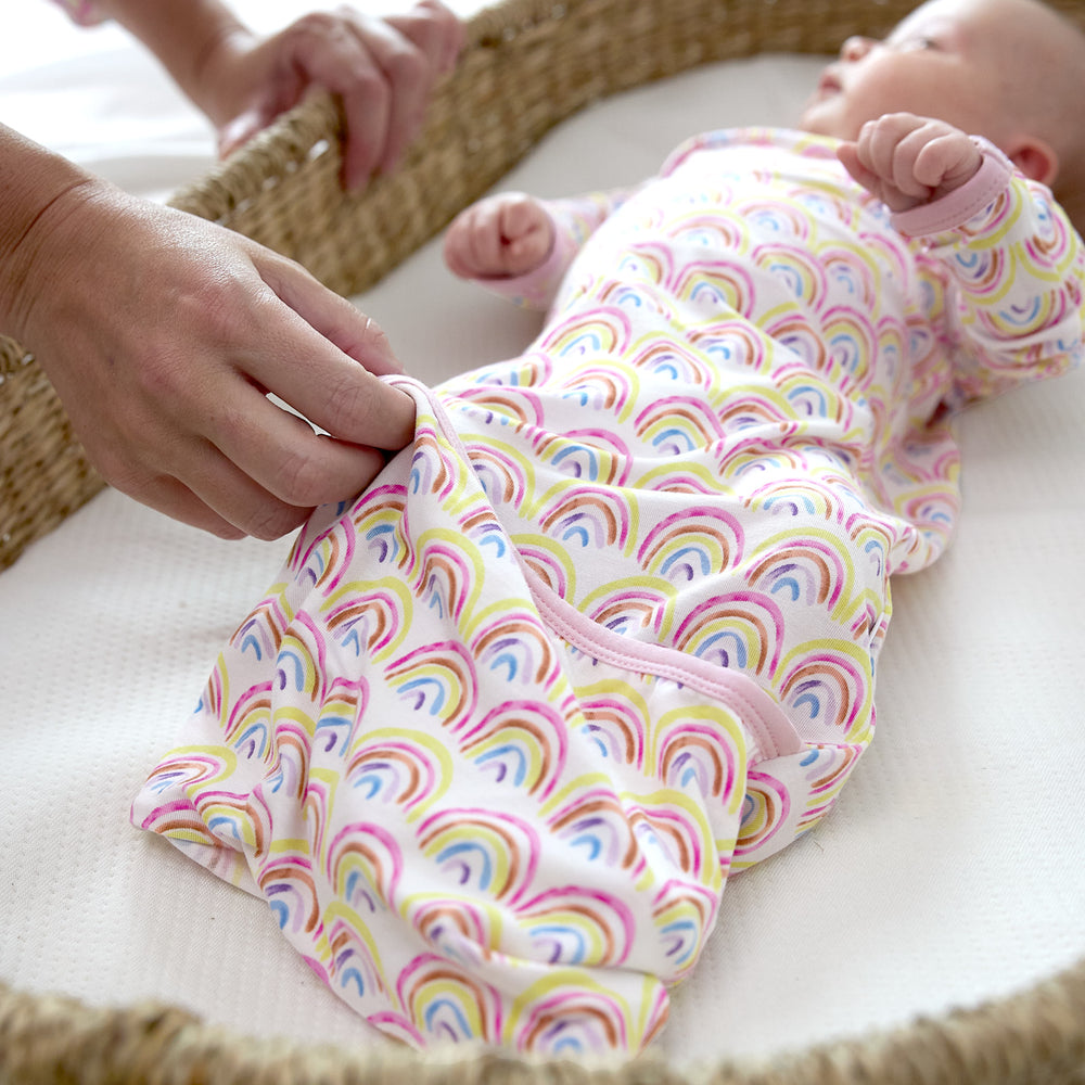 baby in pastel rainbows gown laying down in basket with parents hand folding over