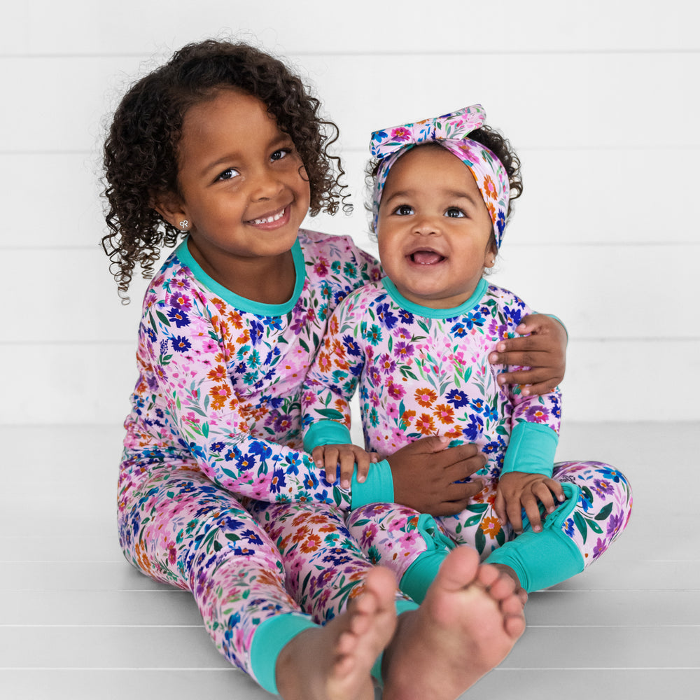 Image of a two children cuddling. one child is wearing a Sweet Pea Floral luxe bow headband and a matching crescent zippy, the other child is wearing a matching Sweet Pea floral two piece pj set