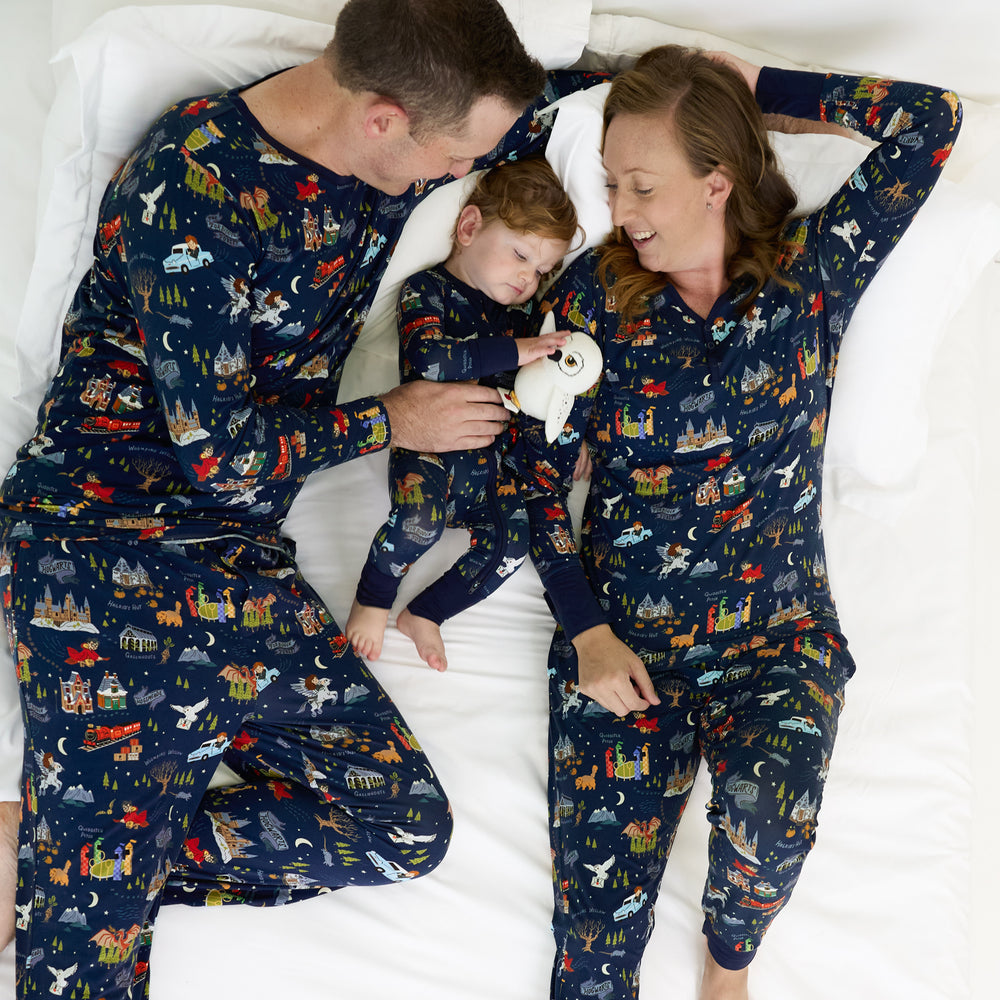 family of three laying on a bed wearing matching An Evening at Hogwarts printed pjs. Dad is wearing a men's pj top and men's pj pants, mom is wearing women's pj top and pj pants, and their child is wearing a zippy and holding a Hedwig Lovey. 