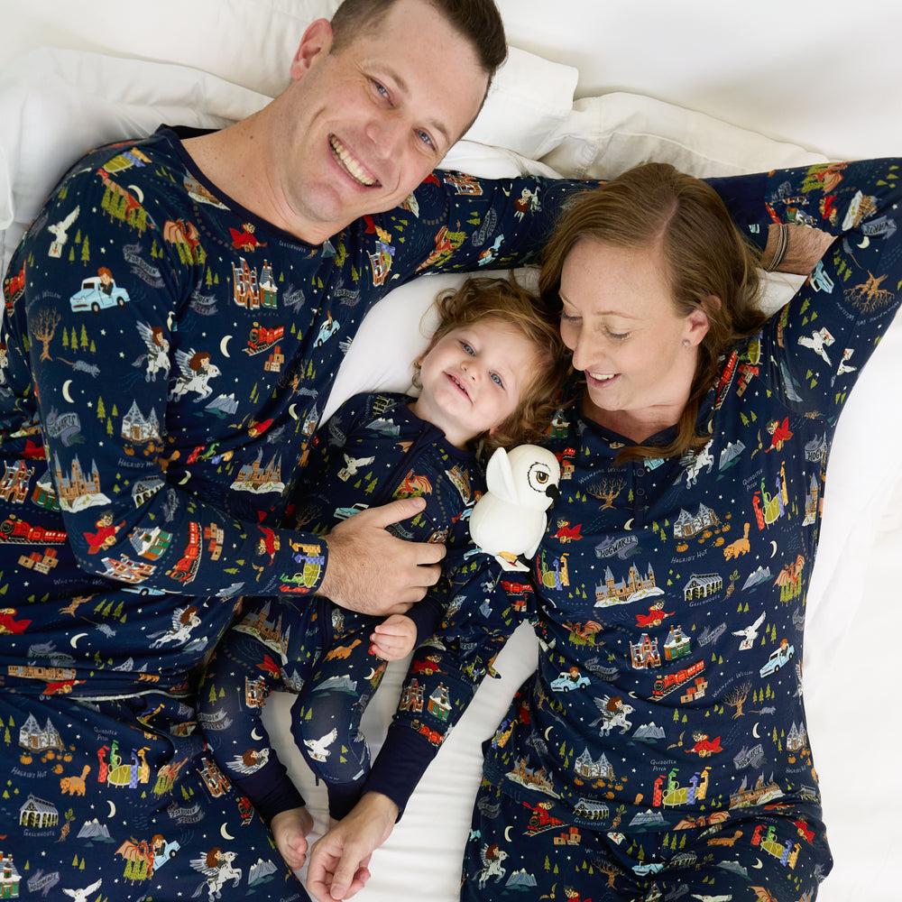 family of three on a bed wearing matching An Evening at Hogwarts printed pjs. Dad is wearing a men's pj top and men's pj pants, mom is wearing women's pj top and pj pants, and their child is wearing a zippy with a Hedwig Lovey