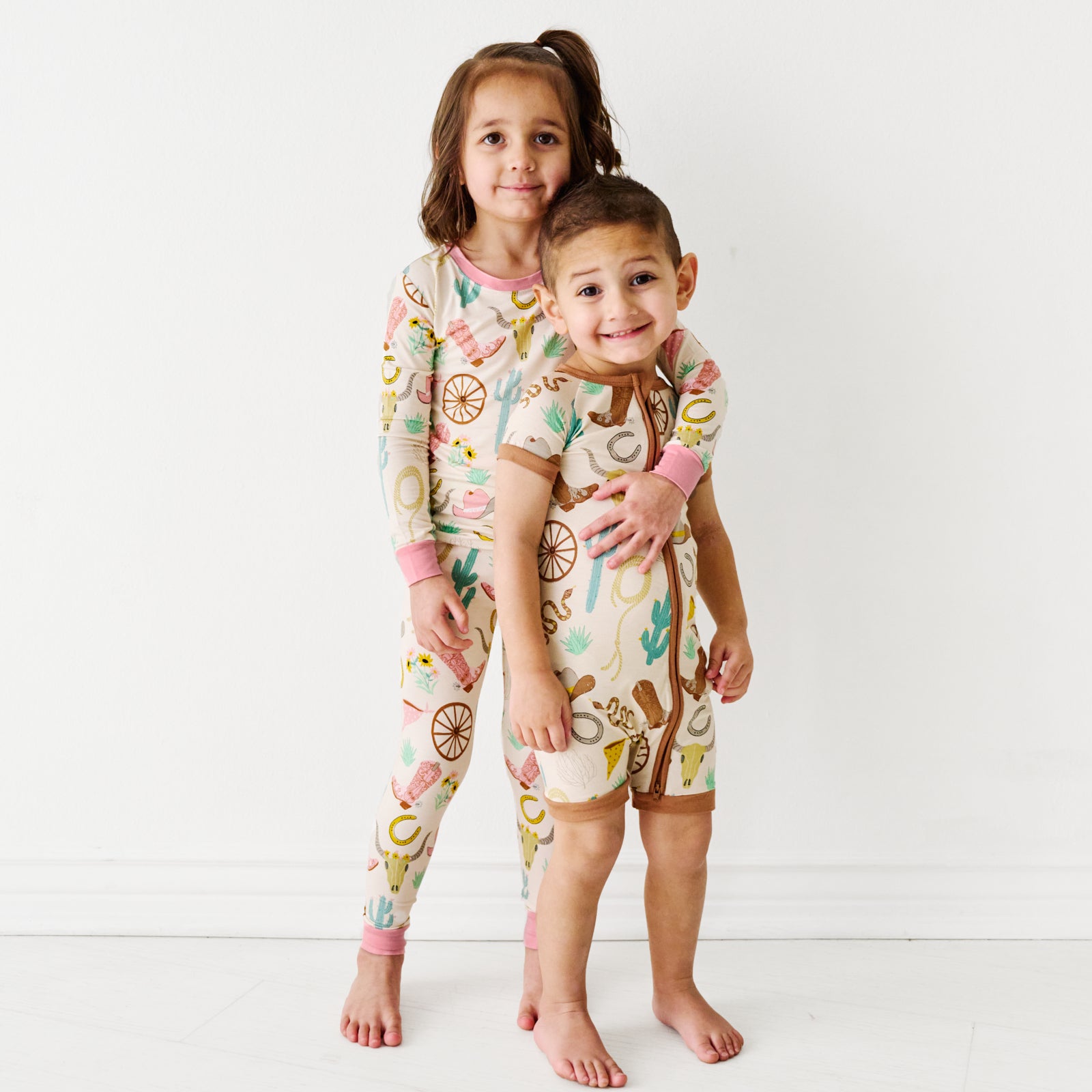 Two siblings posing together wearing coordinating Ready to Rodeo printed pajamas in Pink two piece Ready to Rodeo and Caramel Ready to Rodeo shorty zippy