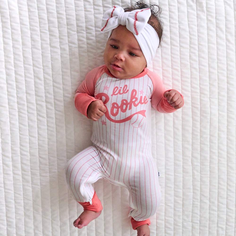 Child laying on a bed wearing a Lil Rookie Crescent zippy paired with a Baseball luxe bow headband