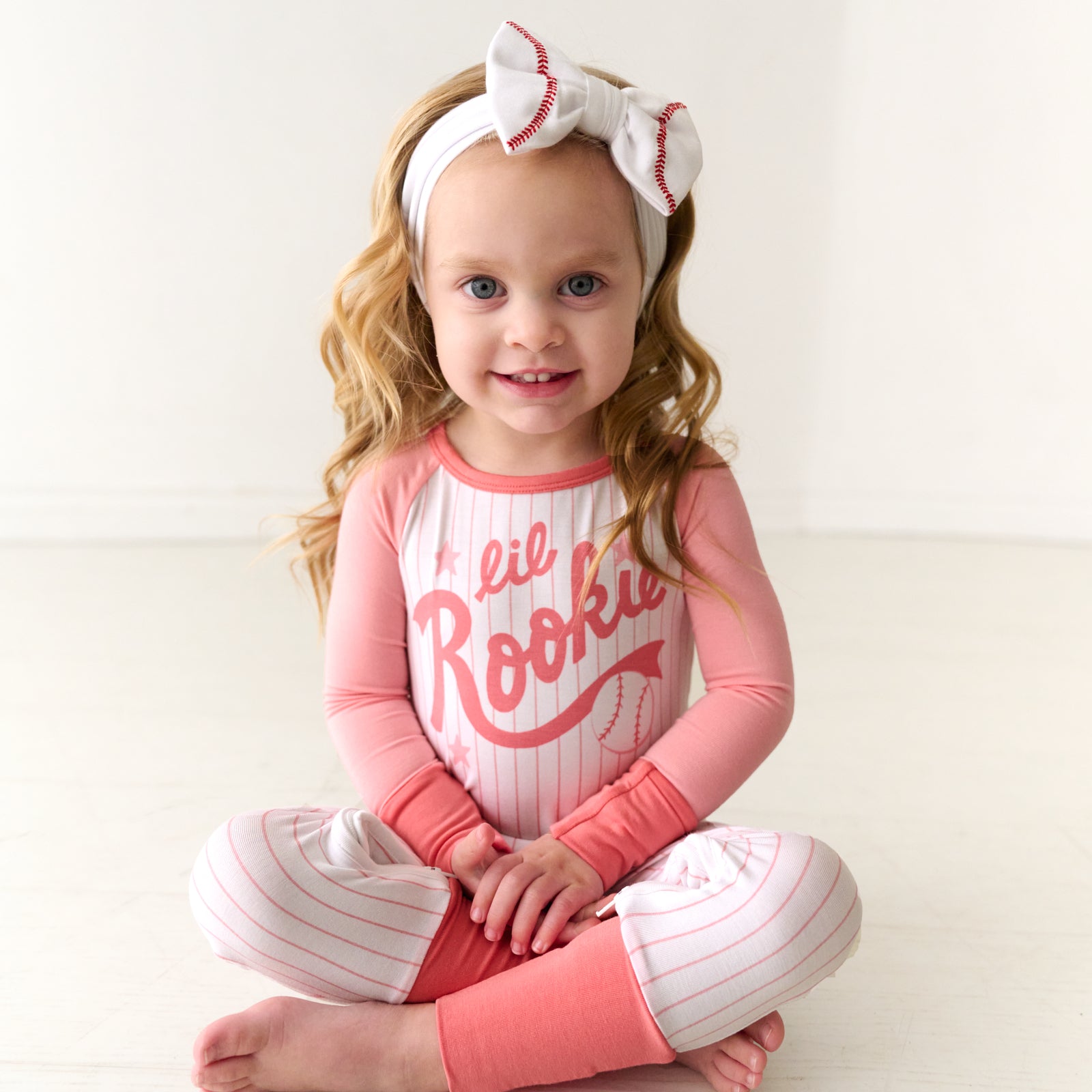 Child sitting wearing a pink Little Rookie crescent zippy paired with a baseball luxe bow headband