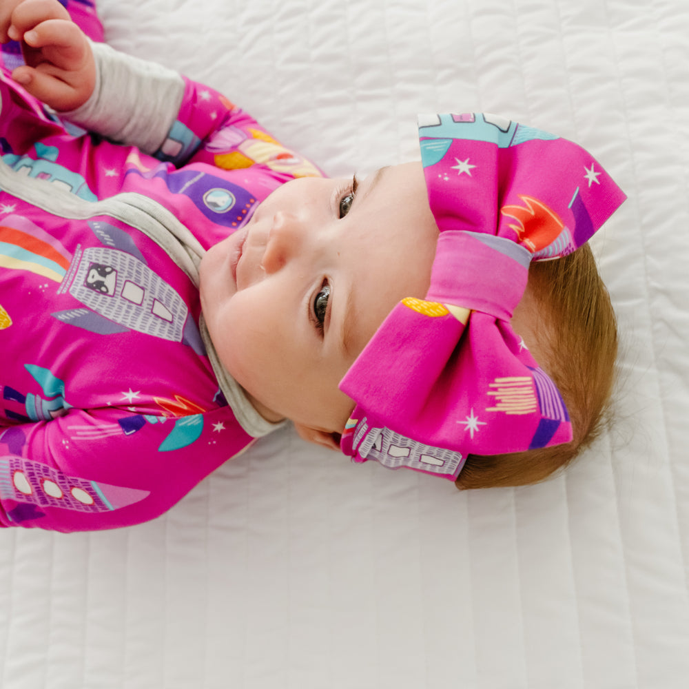 Close up image of a child laying on a bed wearing a Pink Space Explorer zippy paired with a matching luxe bow headband