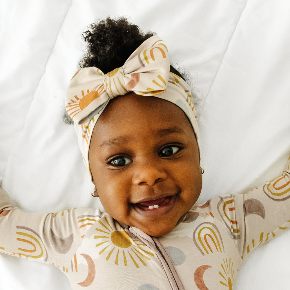 Close up image of a child laying on a bed wearing a Desert Sunrise luxe bow headband and matching zippy