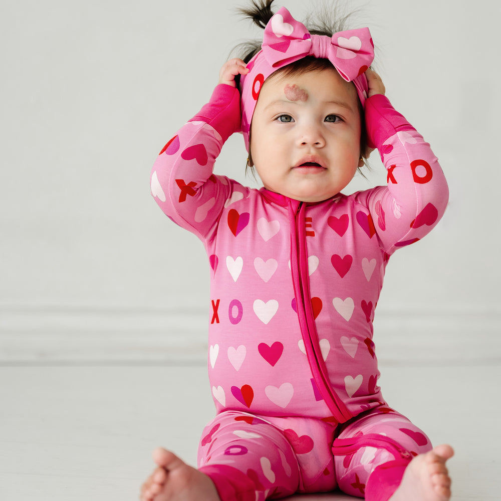 Click to see full screen - Image of a child sitting wearing a Pink XOXO zippy paired with a matching luxe bow headband