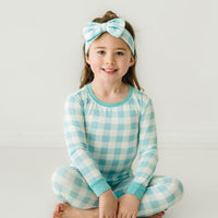 Child sitting wearing an Aqua Gingham two piece pajama set paired with a matching luxe bow headband