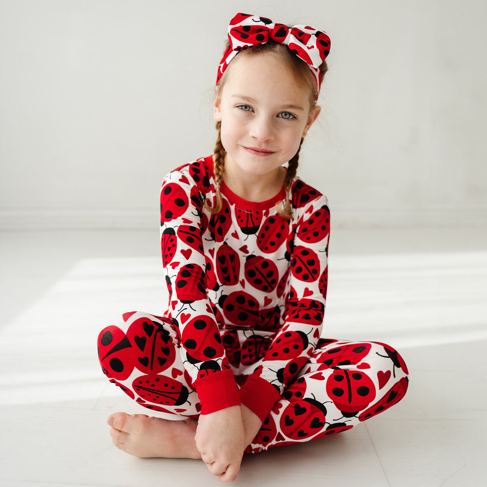 Click to see full screen - Child sitting on the ground wearing a Love Bug printed luxe bow headband and matching pajamas