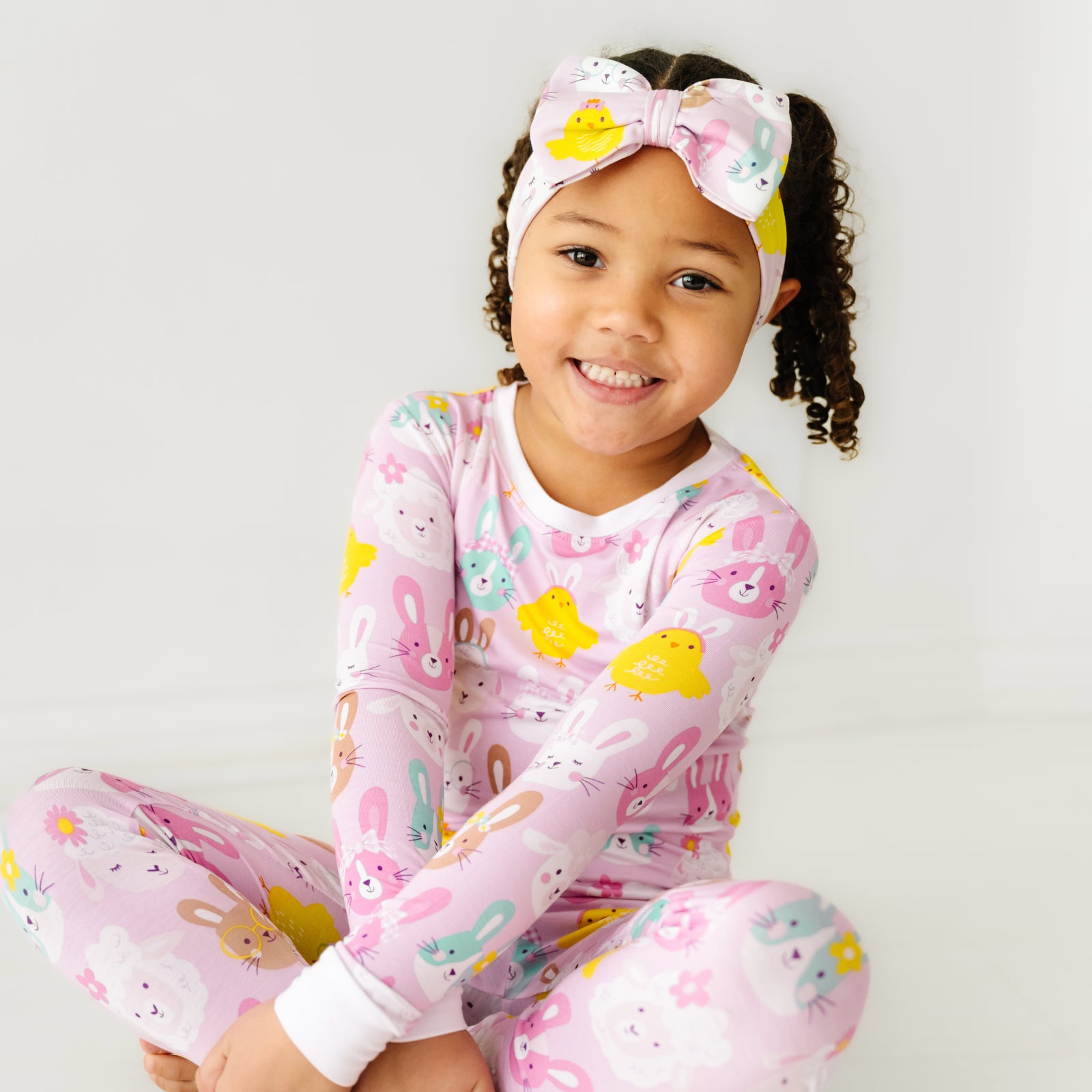  image of a child sitting wearing a Pink Pastel Parade two piece pajama set paired with a matching luxe bow headband