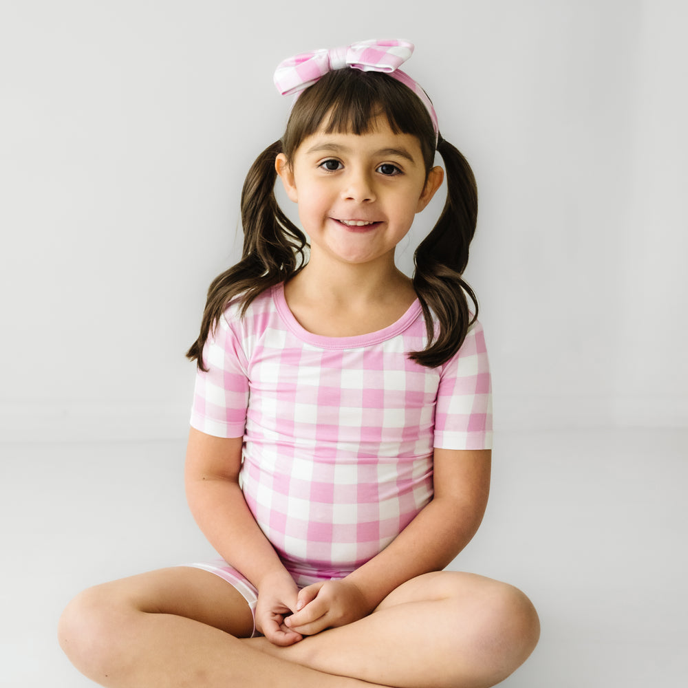 Click to see full screen - child sitting wearing a Pink Gingham two piece short sleeve and shorts pajama set paired with a matching luxe bow headband