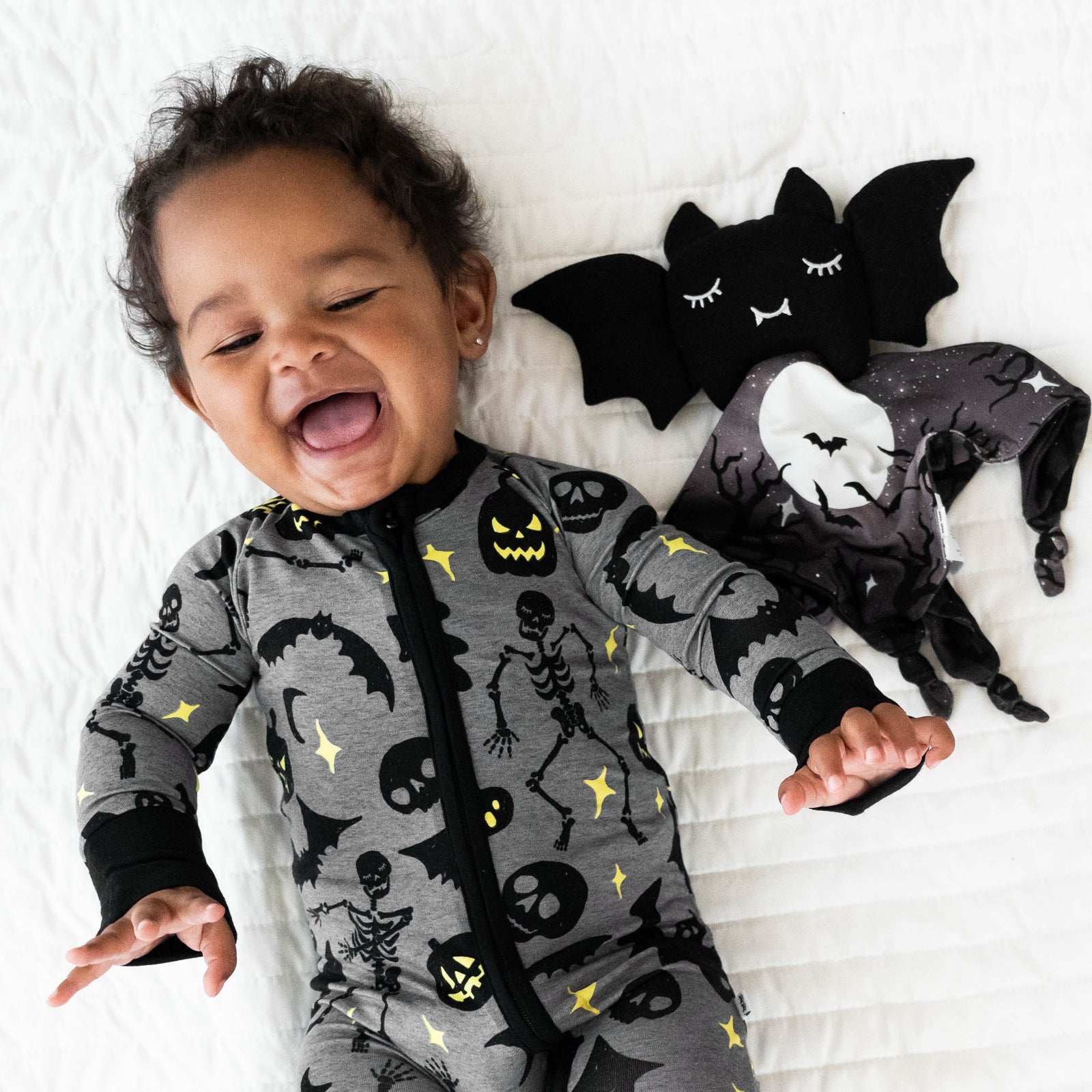 Top view image of baby laying down next to Benny the Bat Sleepyhead Lovey, while wearing the  Gray Glowing Ghouls Zippy 